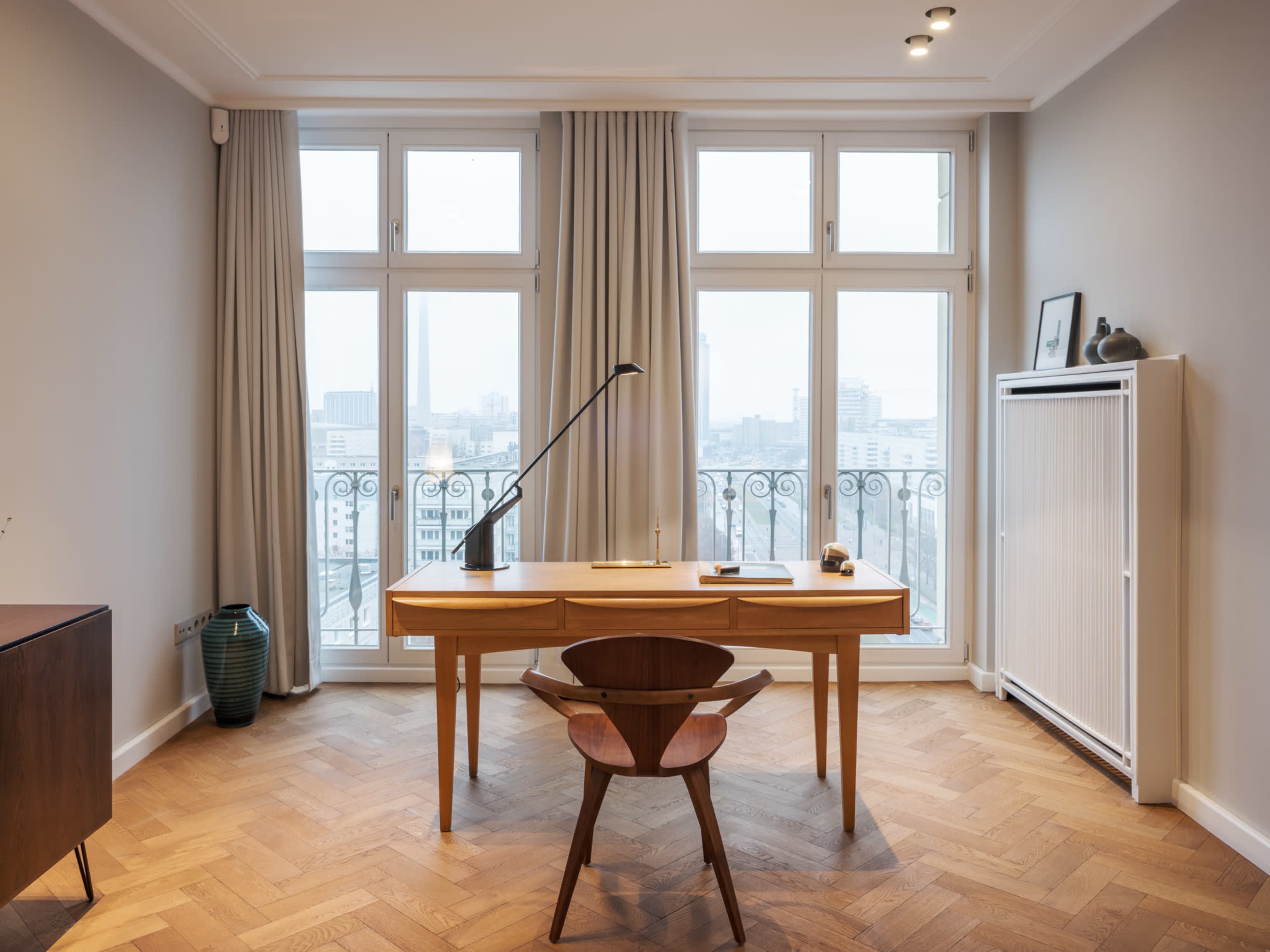 A wooden desk and chair are positioned in front of large windows that overlook a cityscape, with sheer curtains softly framing the view.