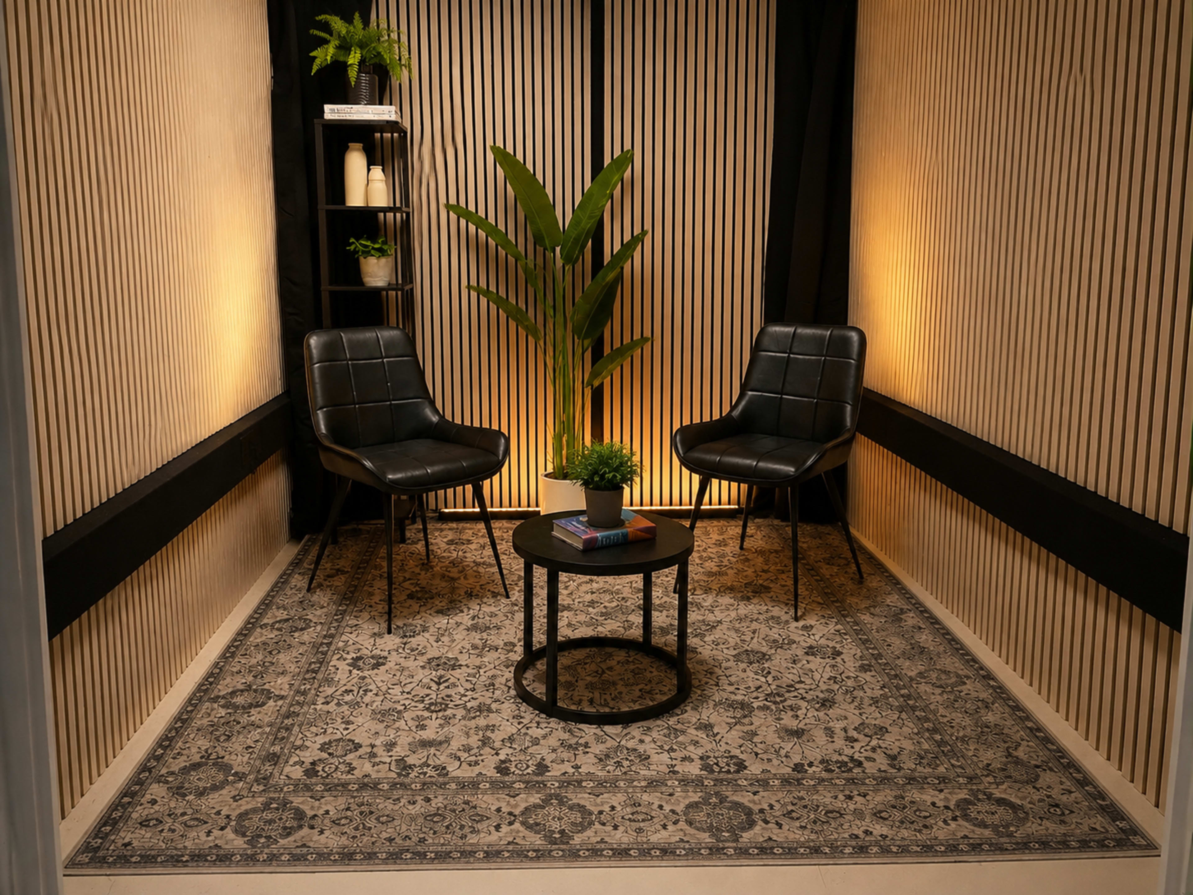 The scene features two black chairs facing each other around a small circular table, set against a backdrop of vertical wooden slats and soft lighting, with a plant and shelves adding decorative elements.