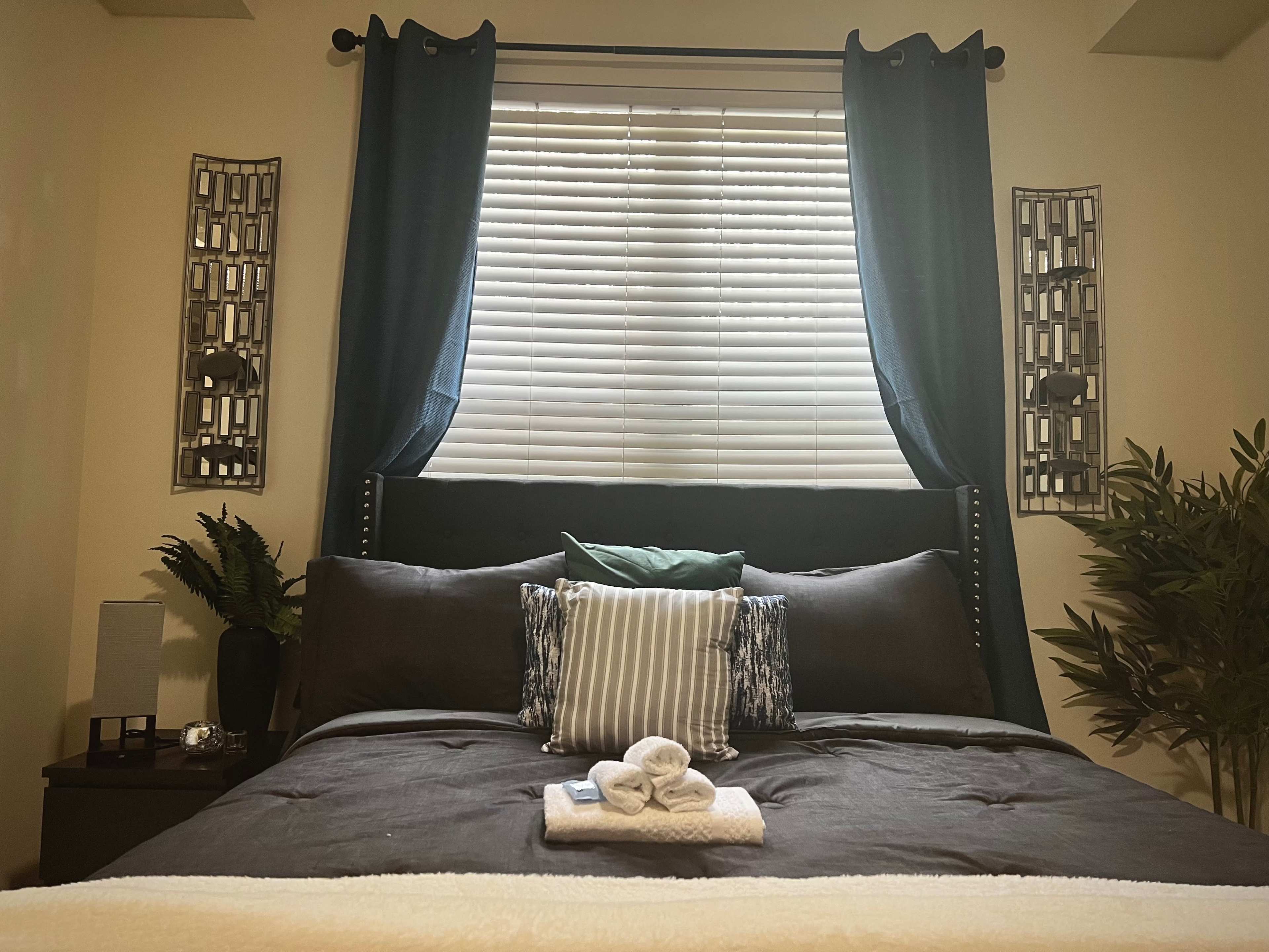 A neatly made bed with dark bedding is positioned against a wall, flanked by two decorative wall pieces and curtains, with a window covered by several blinds.