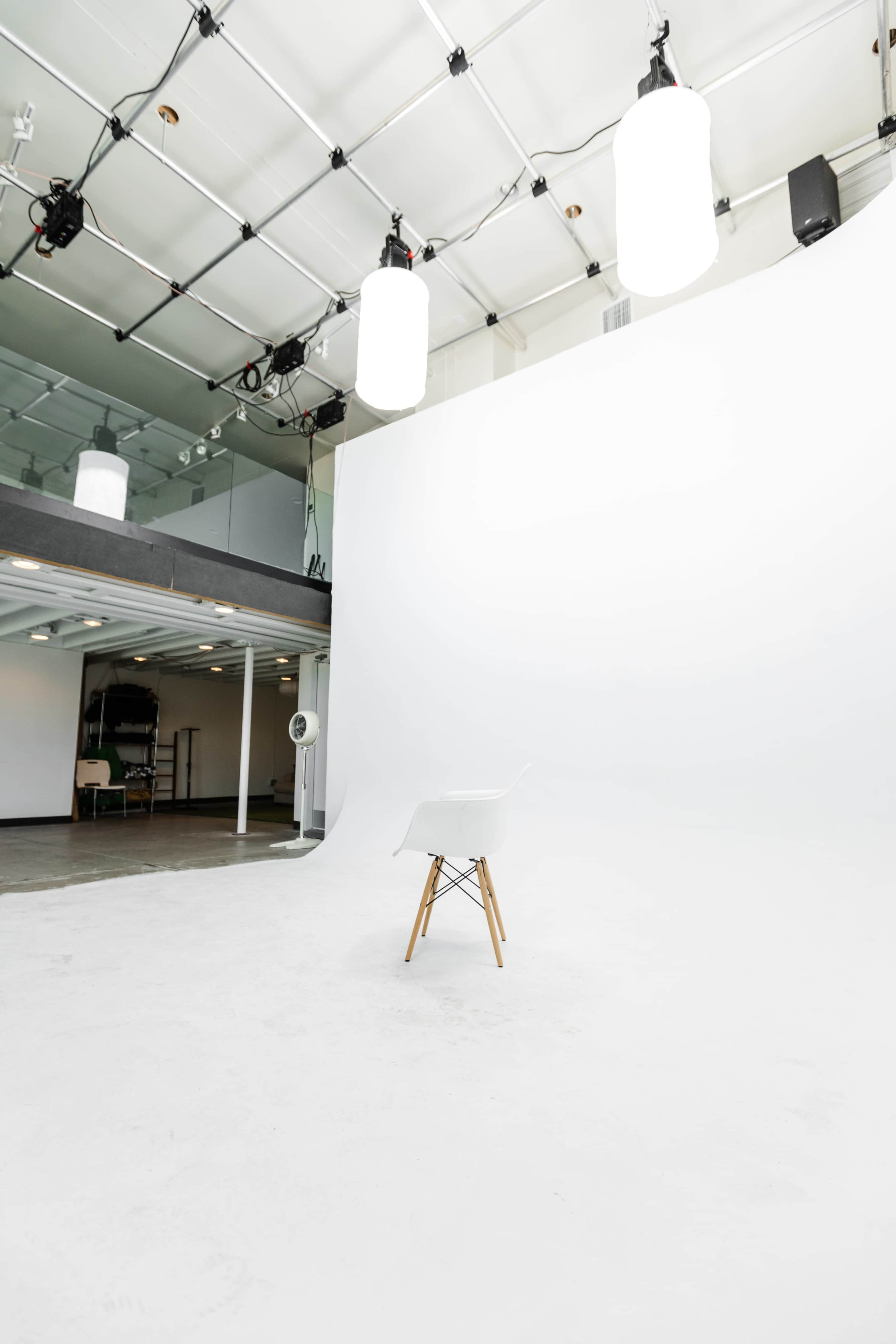 A lone chair is positioned on a smooth white floor beneath soft overhead lighting in an empty studio space.