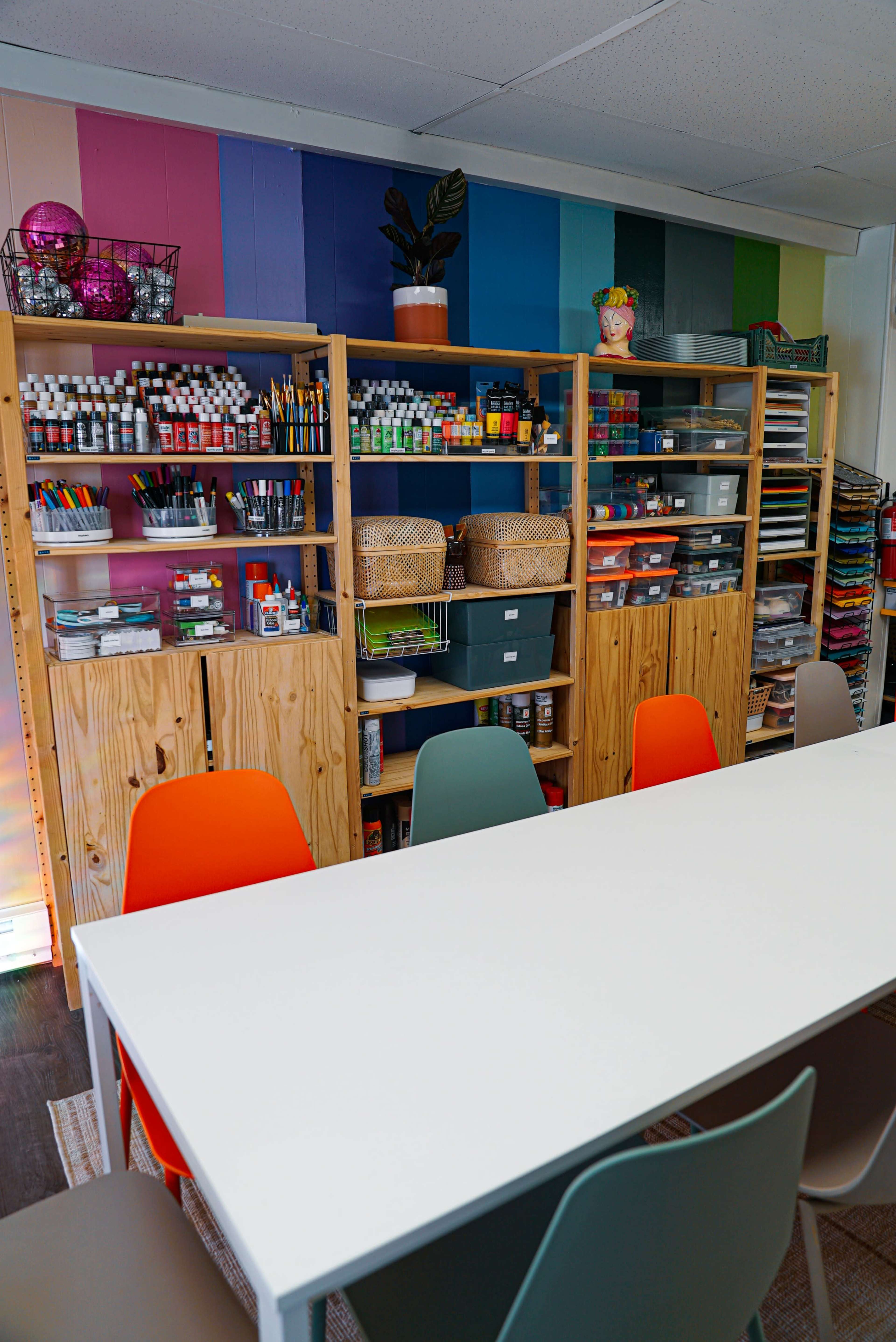 The image shows a brightly colored art studio featuring wooden shelves filled with various art supplies, a long white table, and colorful chairs.