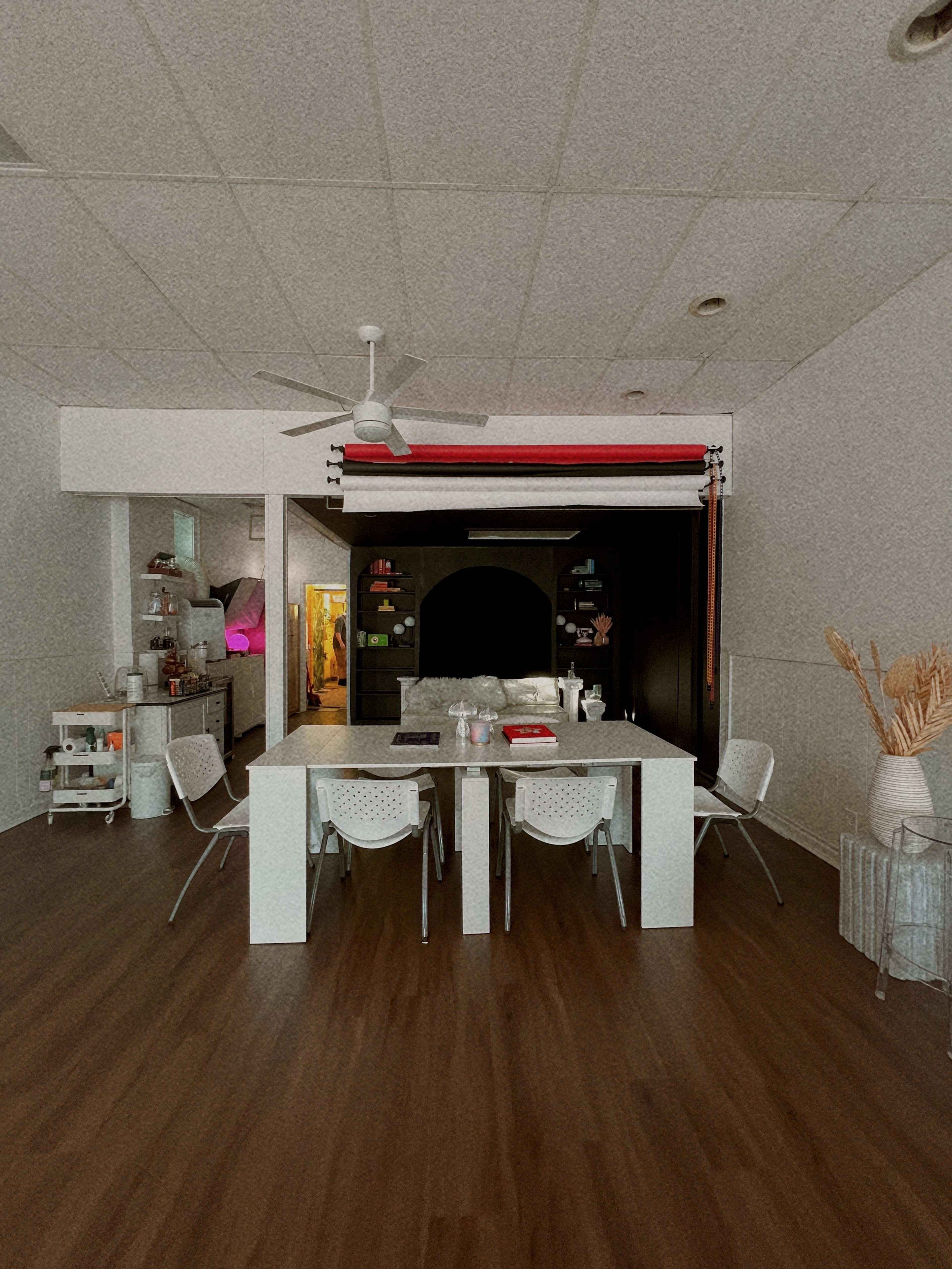 A spacious, well-lit room features a large white table surrounded by chairs, with shelves displaying various items in the background.