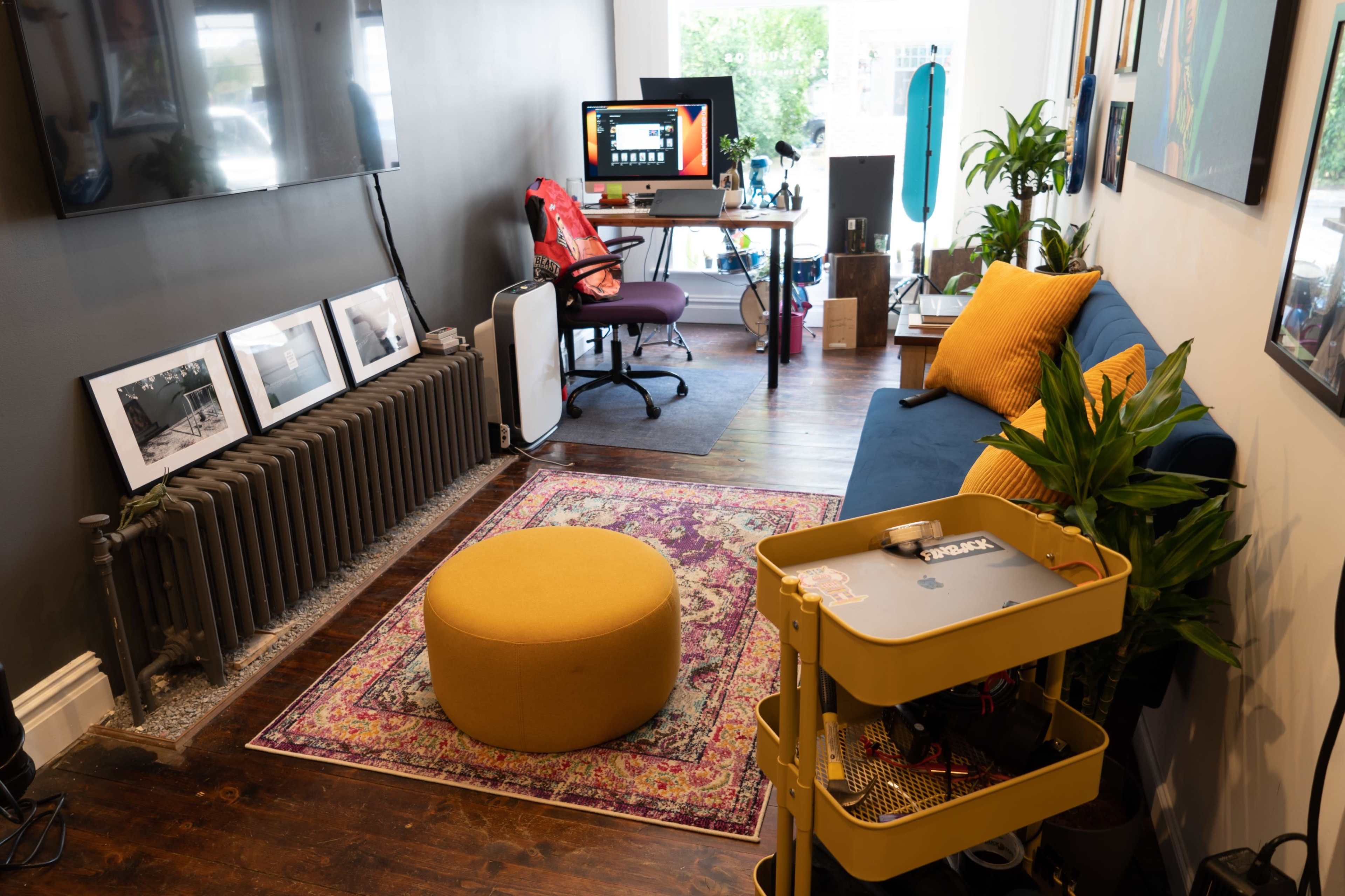 A cozy room featuring a blue sofa, a yellow ottoman, several framed photographs on the wall, a desk with a computer setup, and plants placed throughout the space.