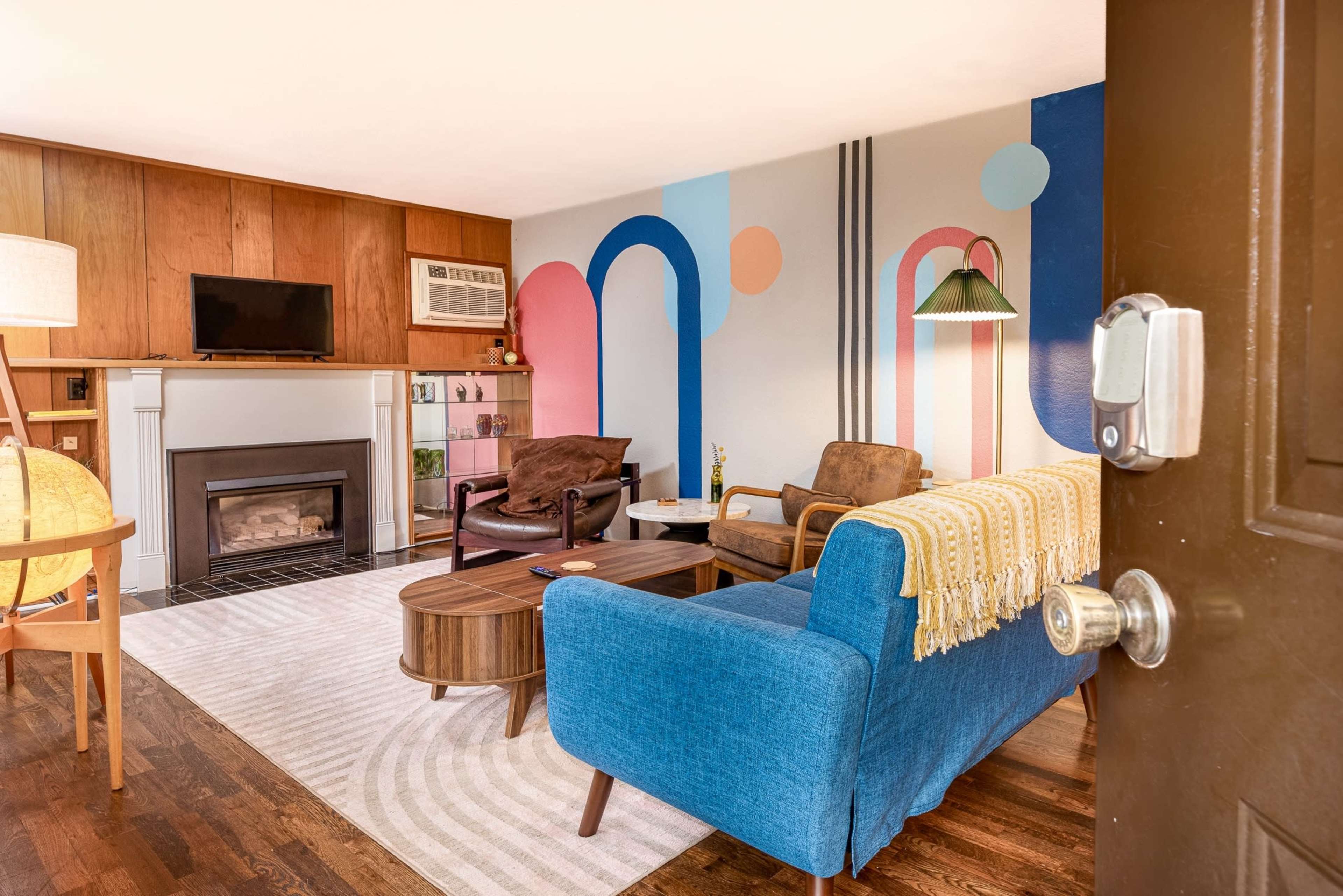 A brightly colored living room features a blue sofa, a coffee table, and a fireplace, with large abstract shapes painted on the wall.