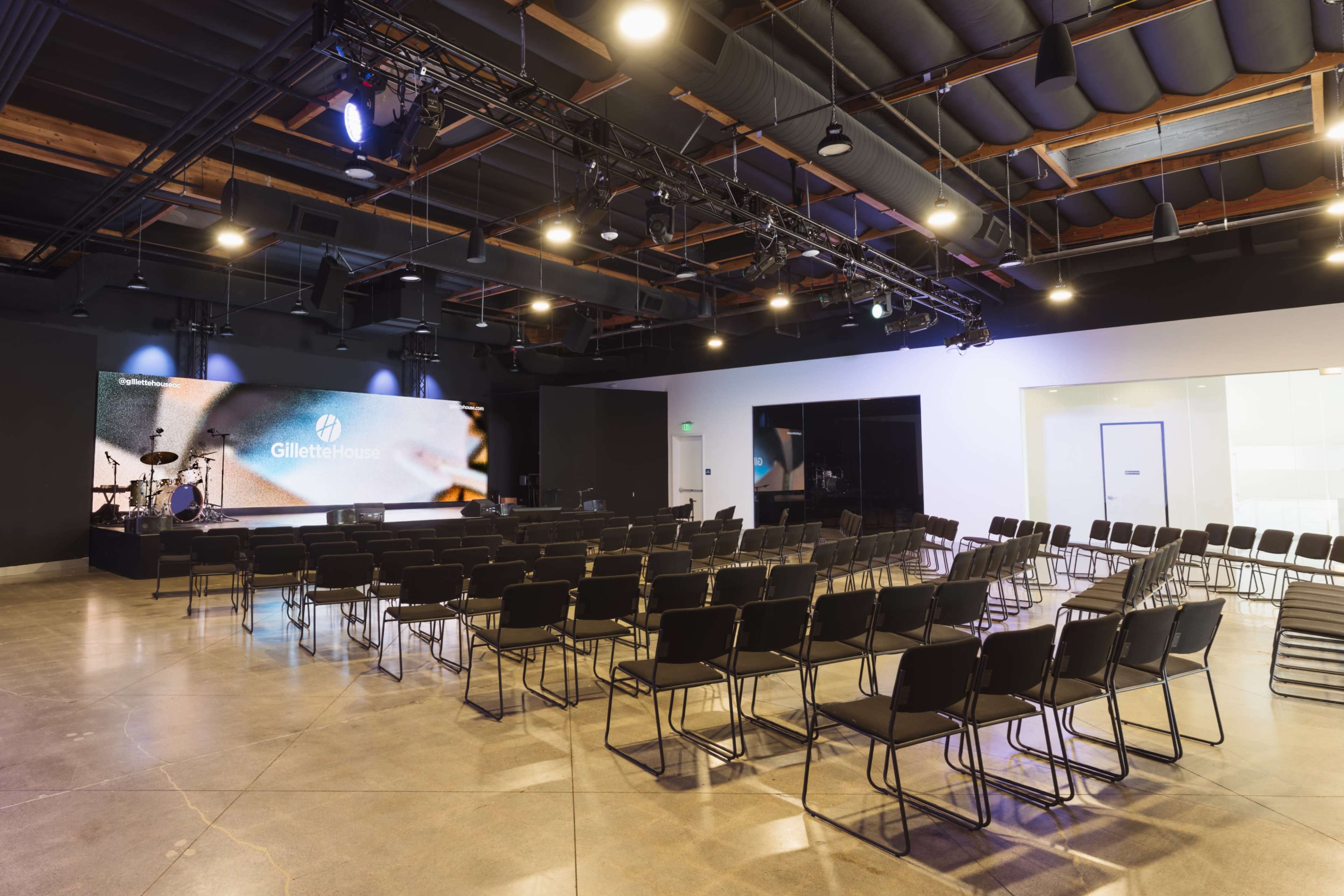 The image shows a modern event space set up with rows of black chairs facing a stage equipped with lighting and screens.