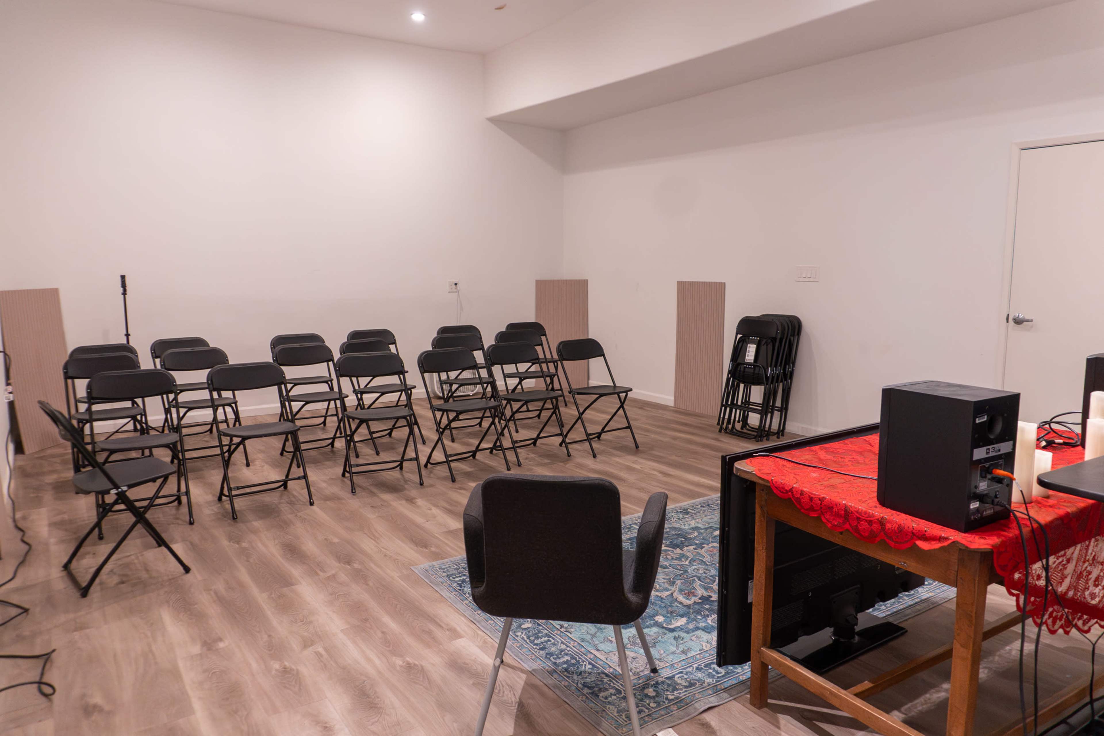 The image shows a sparsely furnished room with several folding chairs arranged facing a table and a chair, along with a rug on the floor.