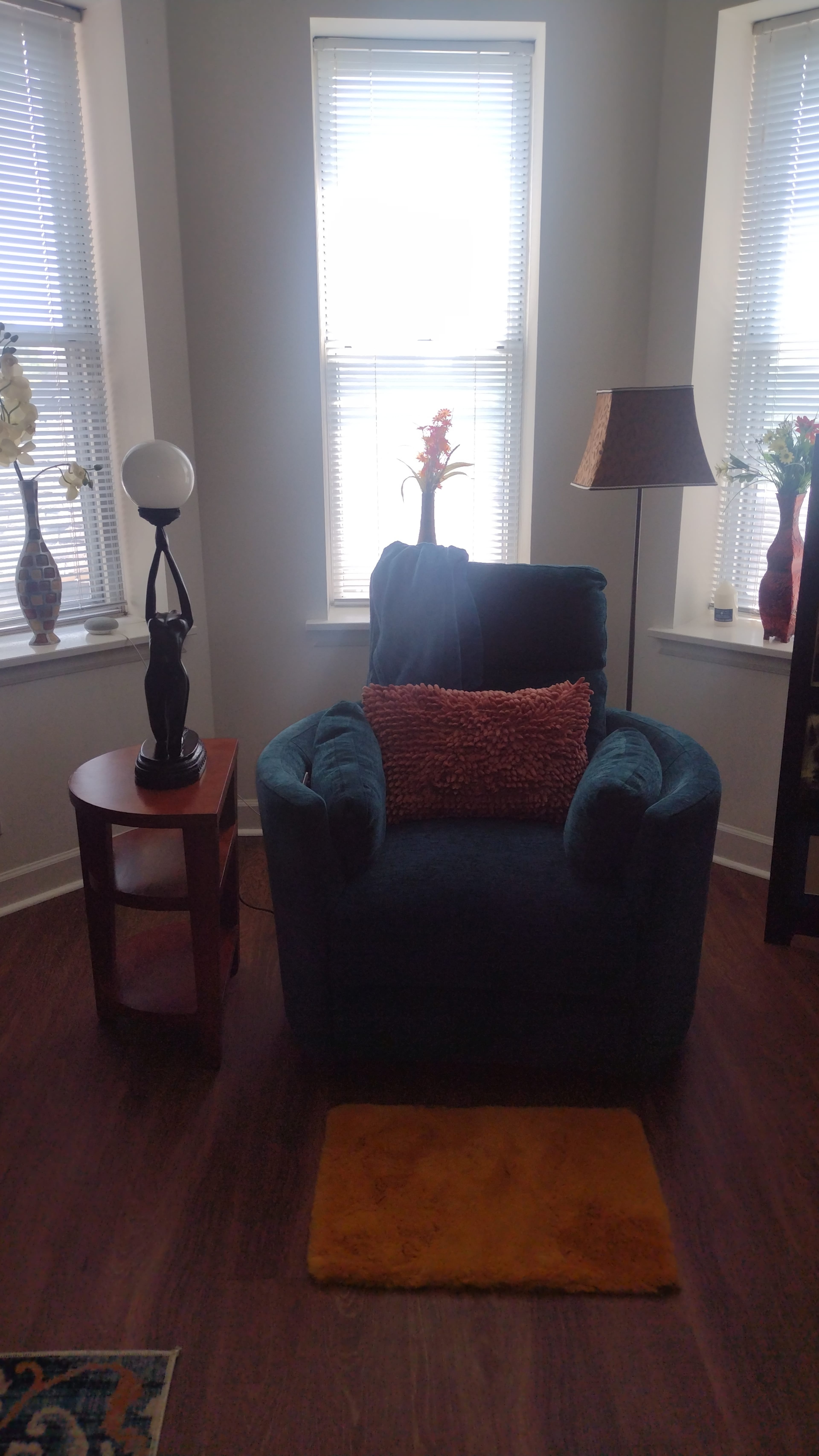 A blue armchair with a decorative orange pillow sits next to a small round table and a lamp in a bright room with two windows.