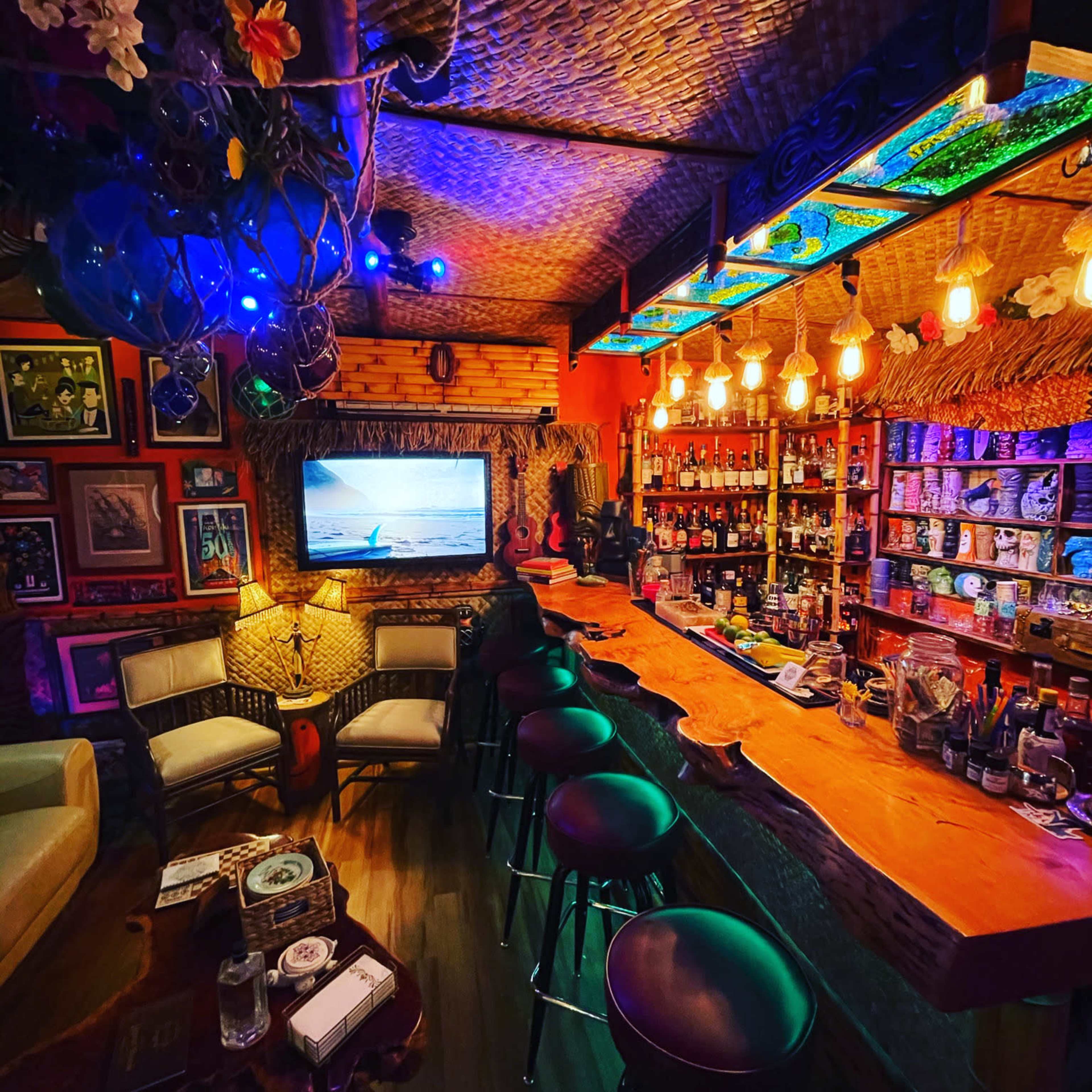 The image shows a cozy tiki bar with a thatched roof, colorful lighting, a wooden bar counter, and shelves stocked with various bottles.