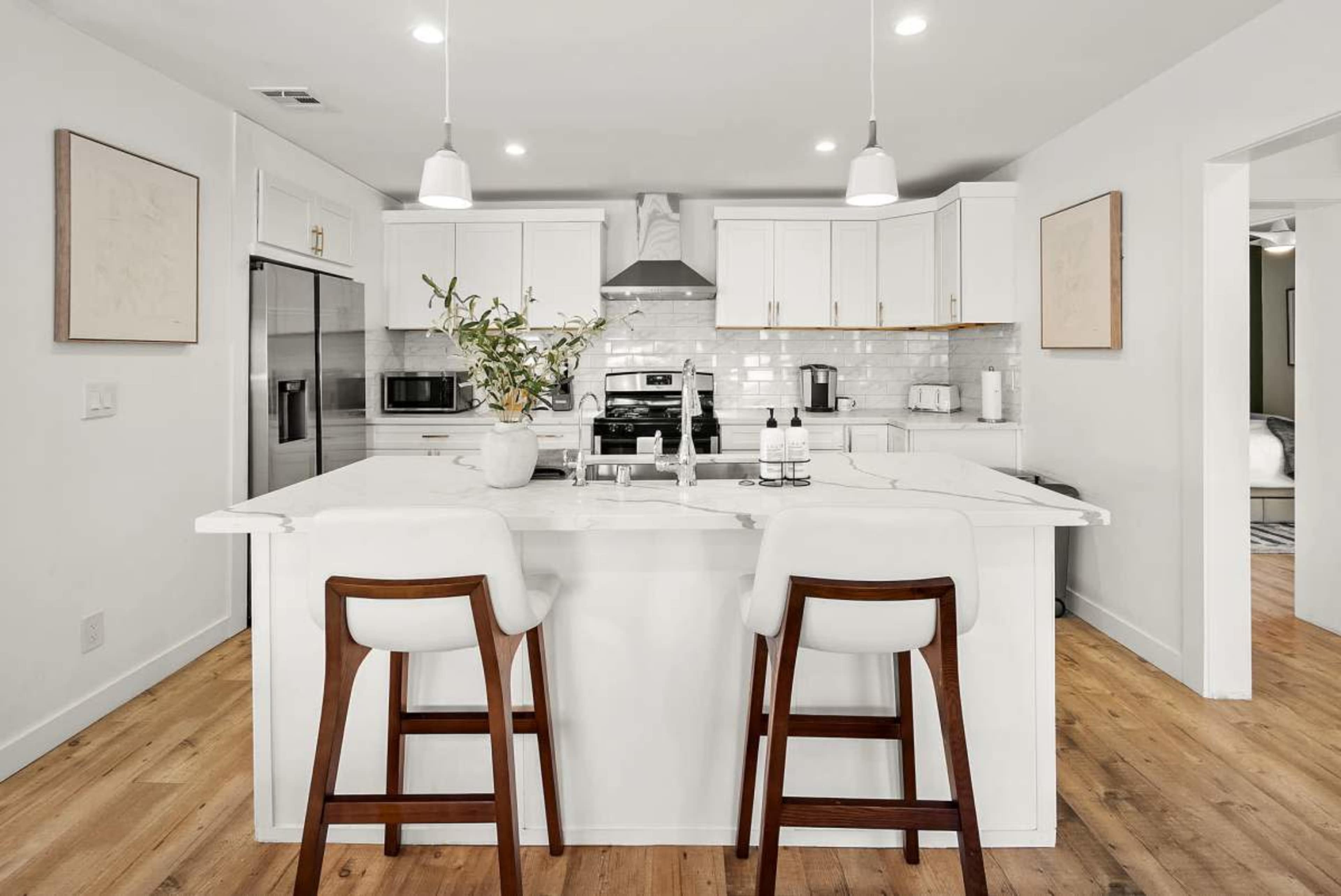 The image shows a modern kitchen with white cabinetry, a central island with bar stools, and stainless steel appliances.