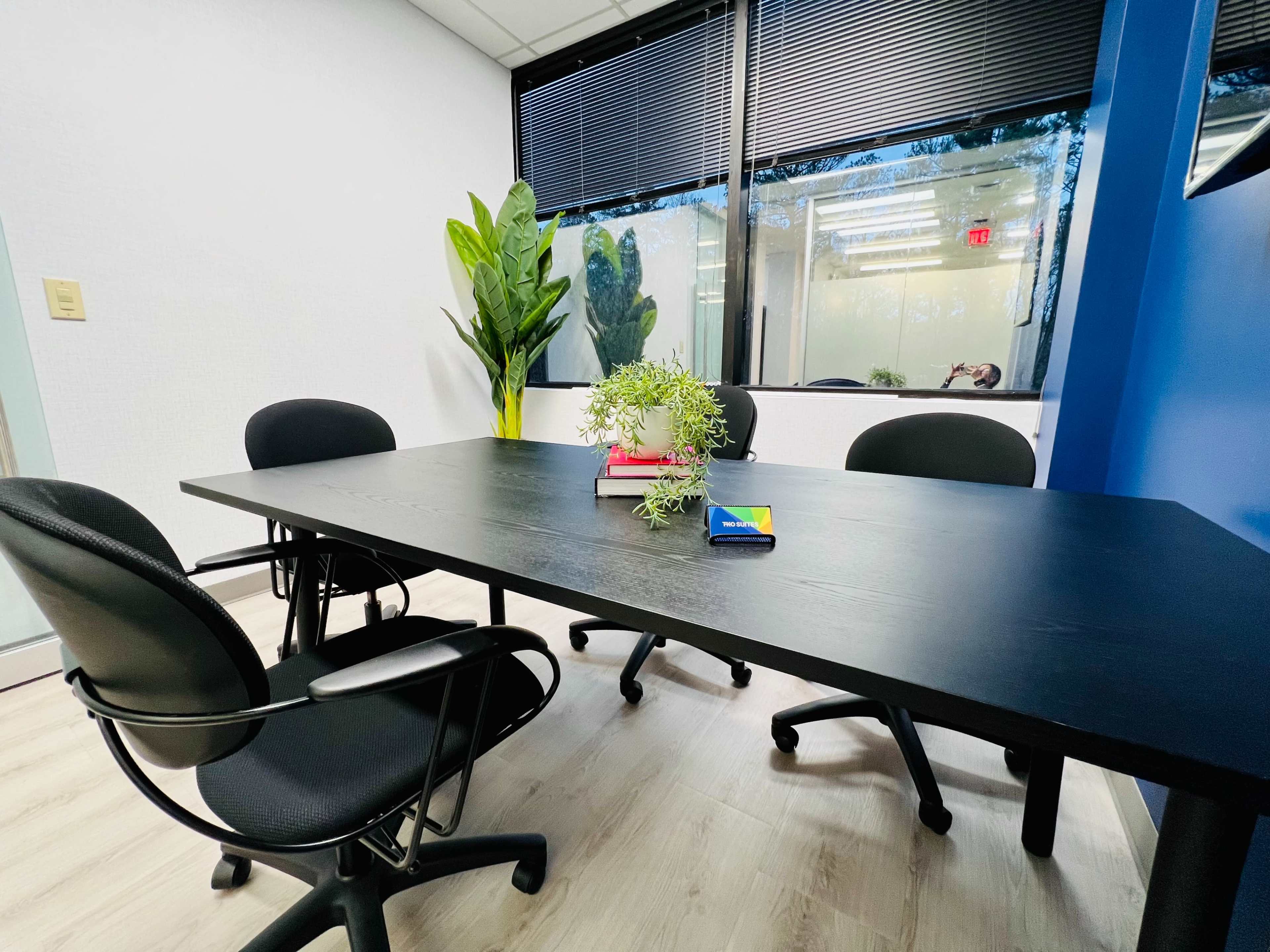 A black table with four chairs is set inside a well-lit conference room featuring a plant and a stack of books at its center.