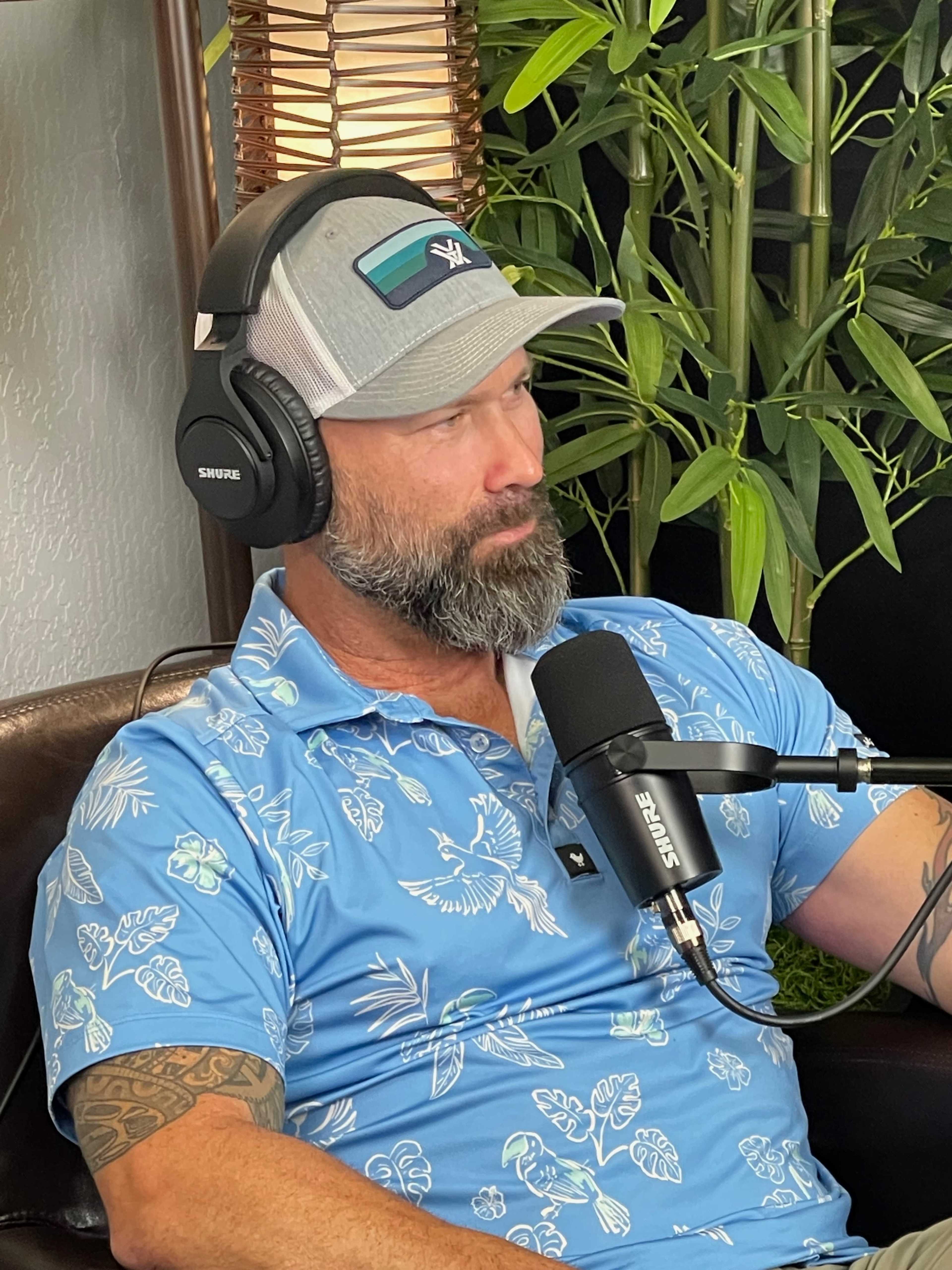A man with a beard is sitting on a couch wearing a blue floral shirt and headphones while speaking into a microphone.