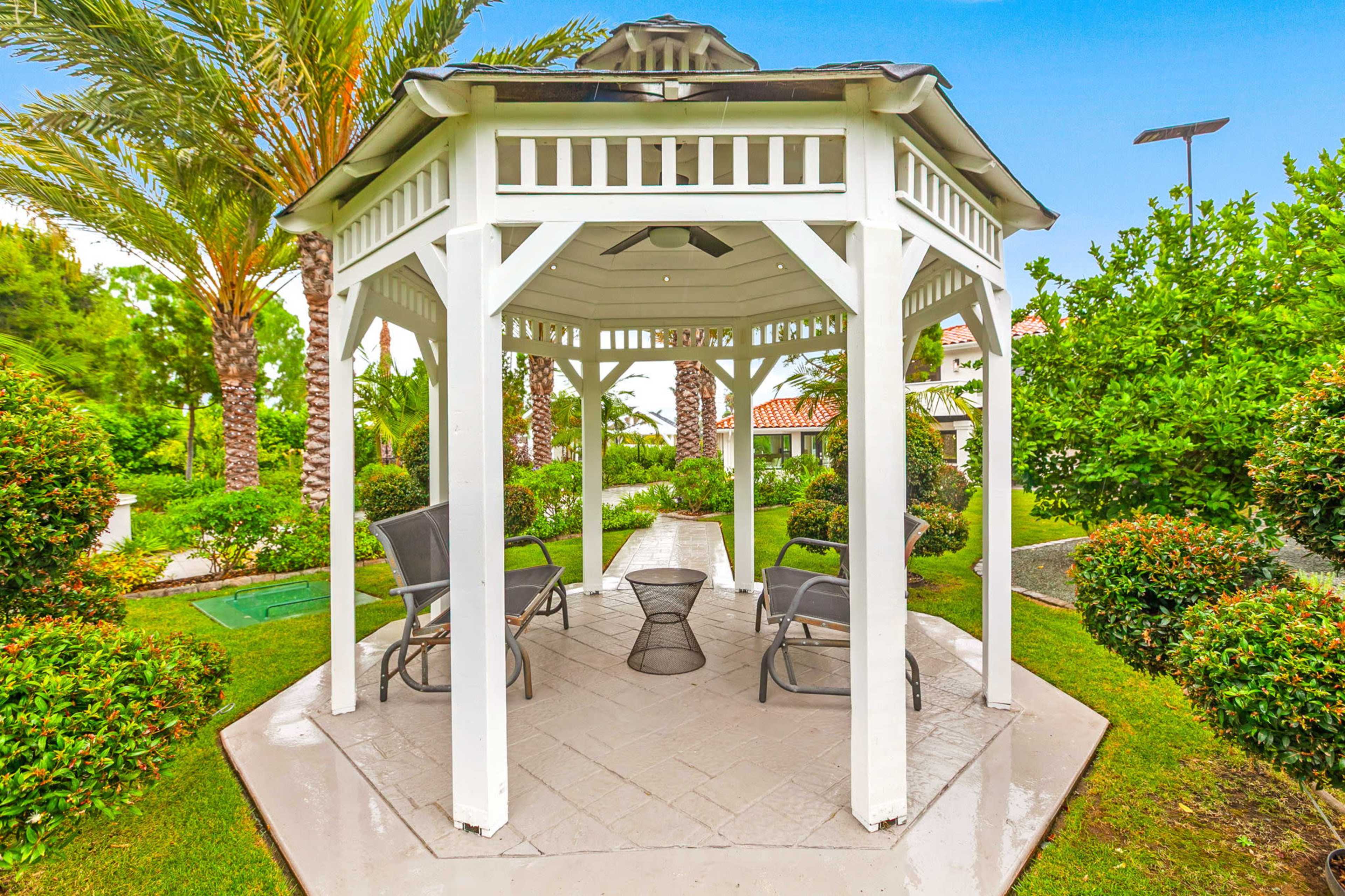 A white gazebo with a tiled floor and metal chairs is surrounded by palm trees and neatly trimmed hedges in a garden setting.