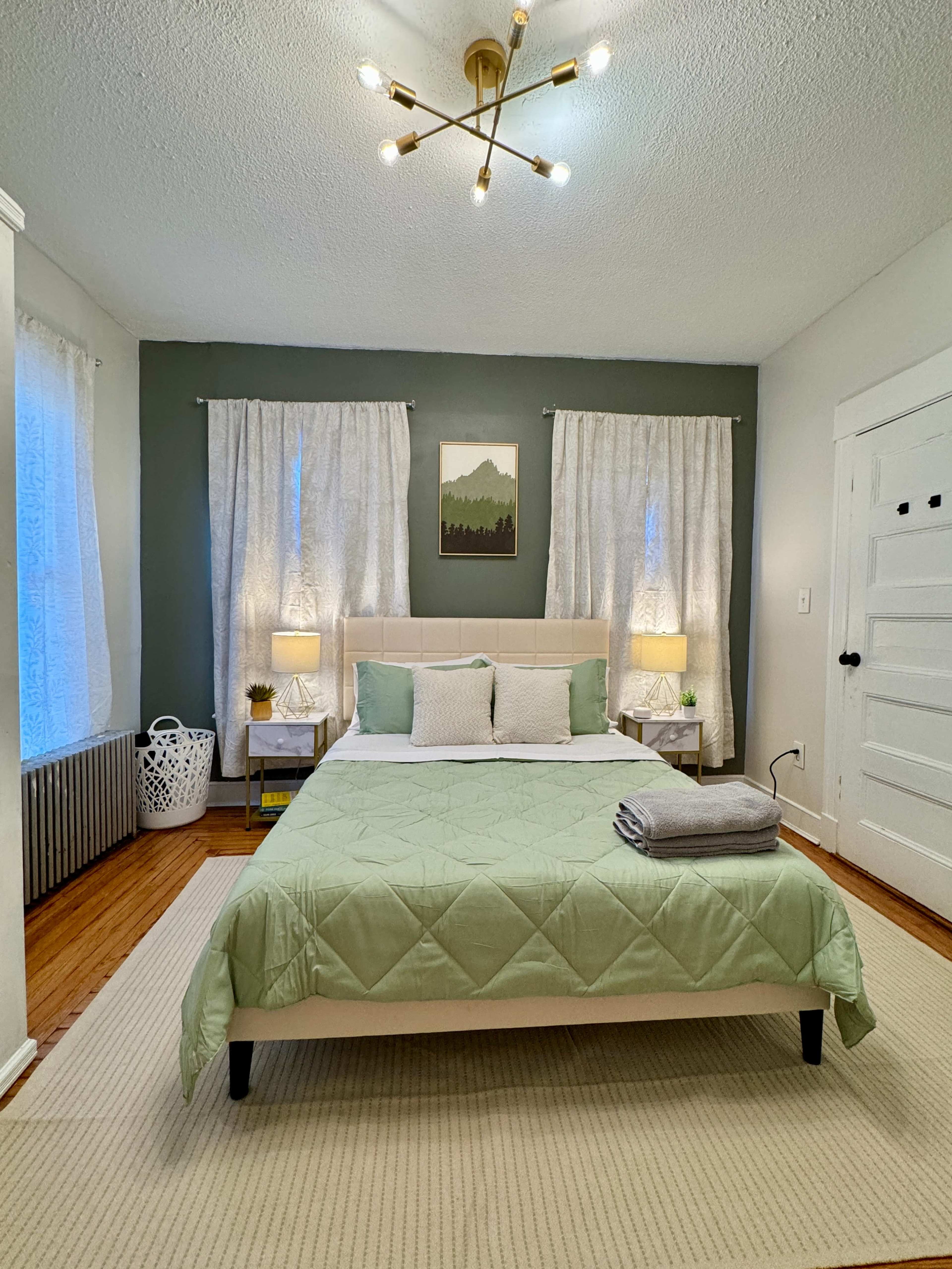 The bedroom features a central bed with a green quilt, two bedside lamps, and light curtains on the windows, complemented by a mountain landscape painting on the wall.