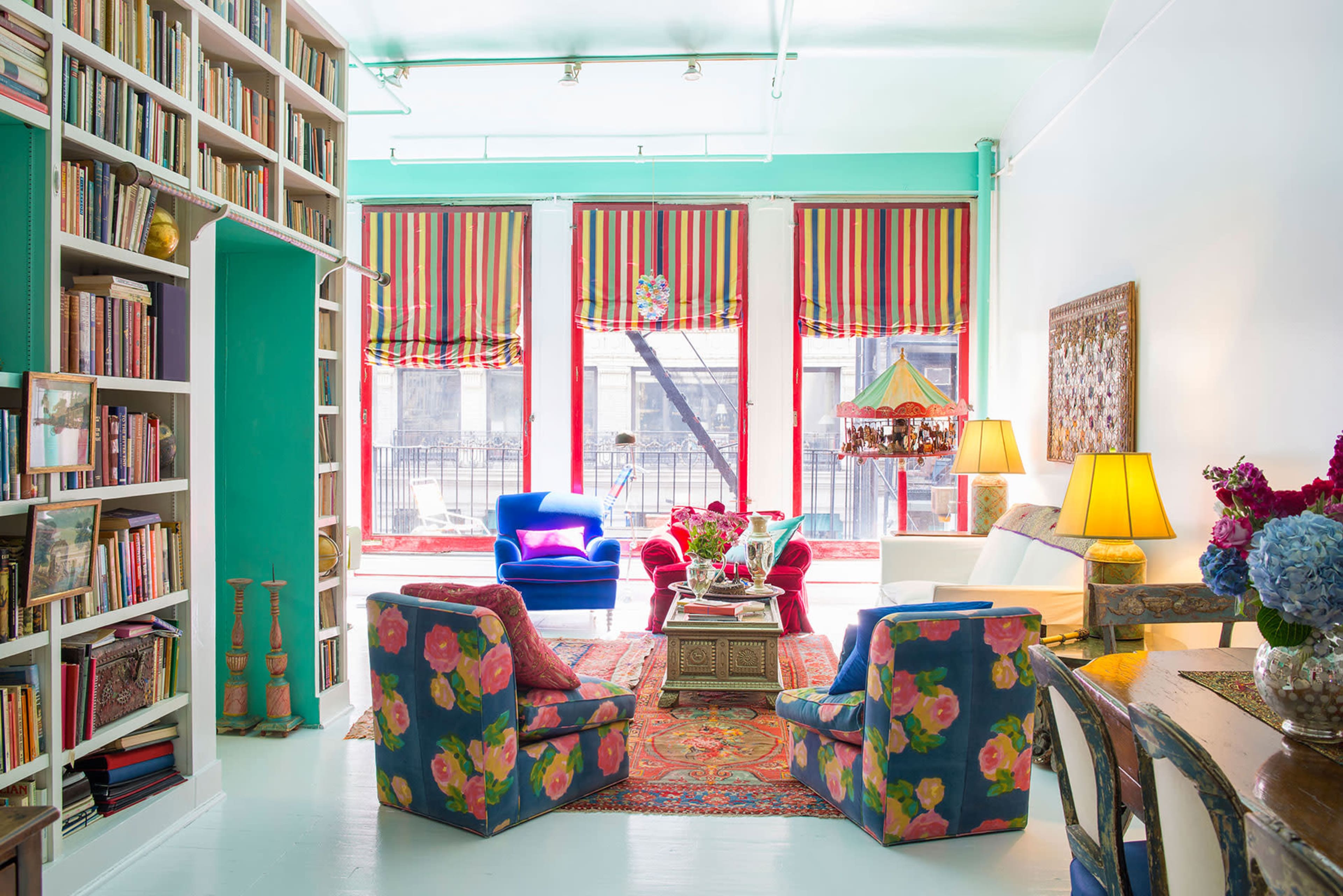 The image shows a brightly colored living room with a bookshelf, colorful upholstered chairs, and large windows with striped curtains.