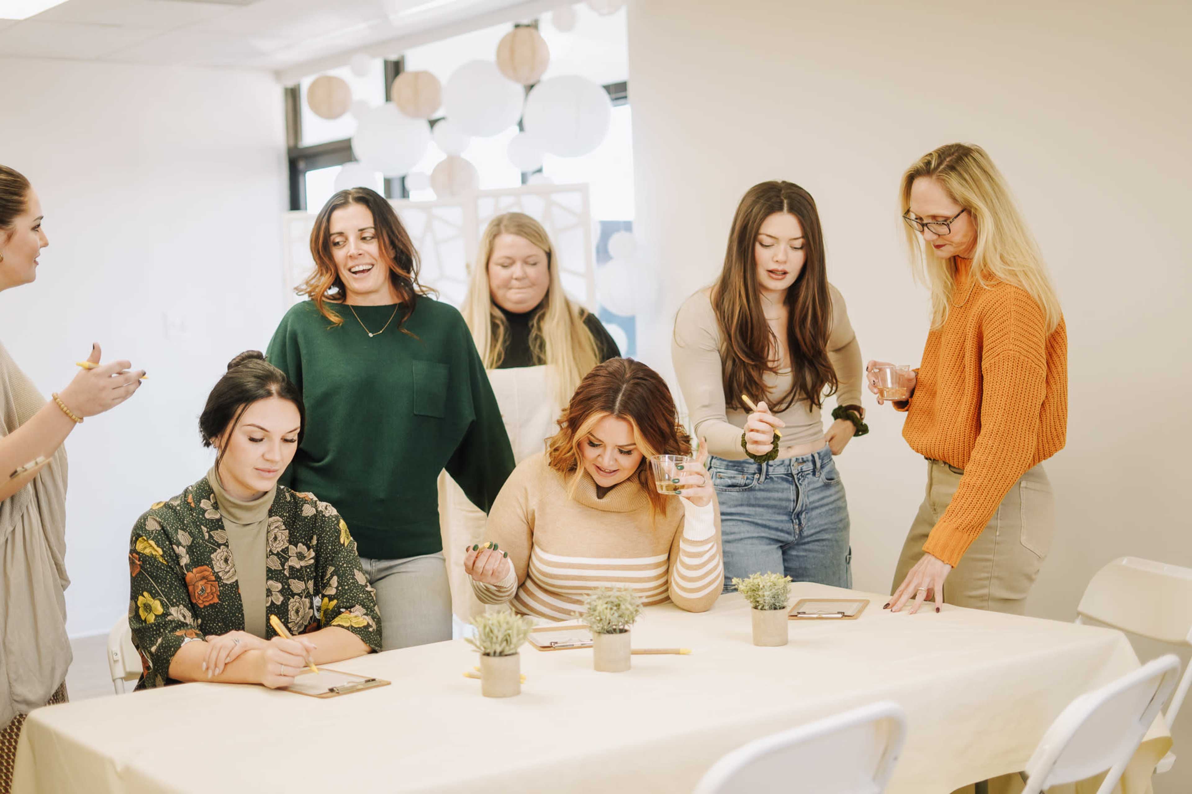 A group of seven women engages in a collaborative activity at a table decorated with small plants, surrounded by soft, illuminated decor.