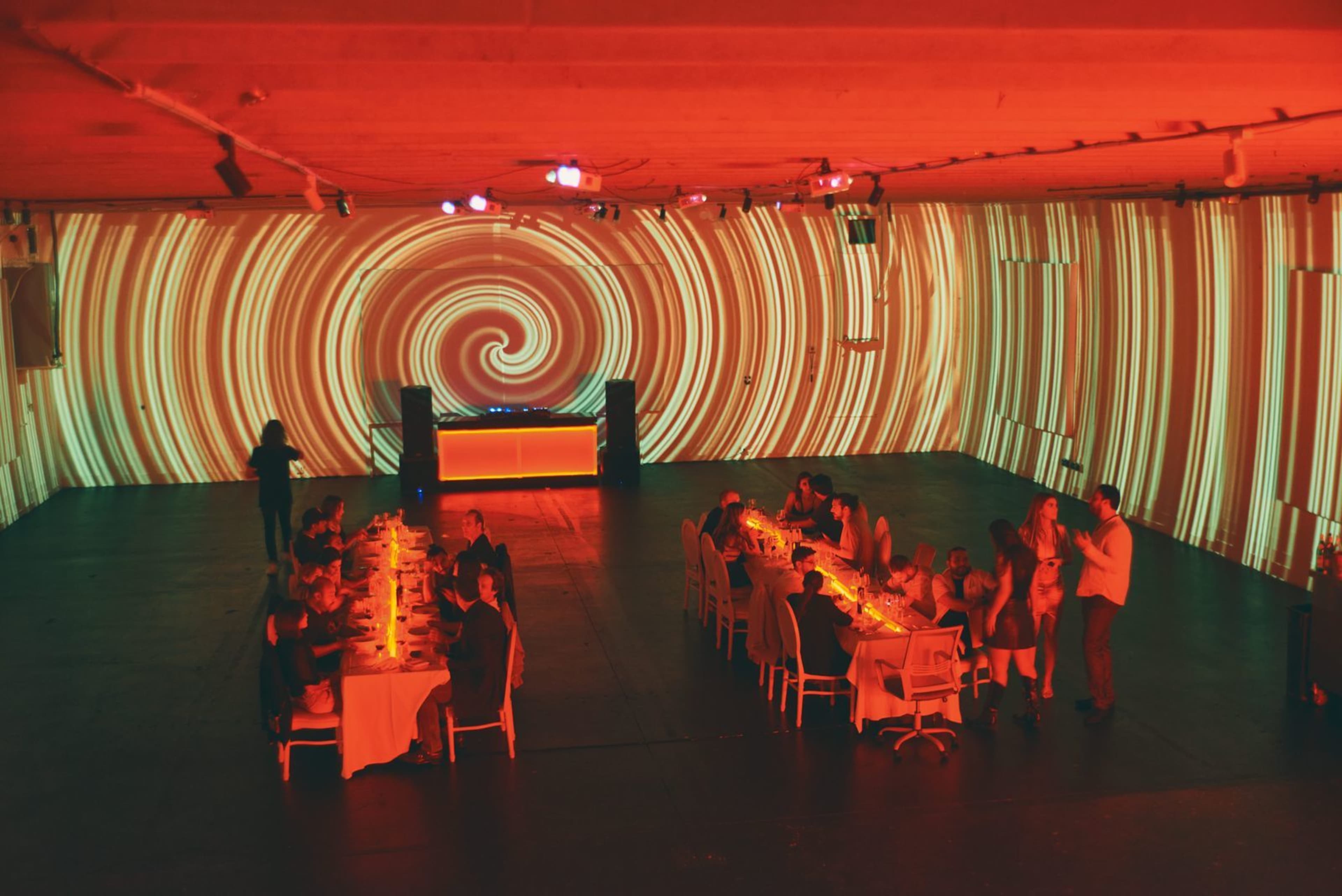 A group of people is gathered around a long table in a dimly lit room with swirling red and orange light projections on the walls.