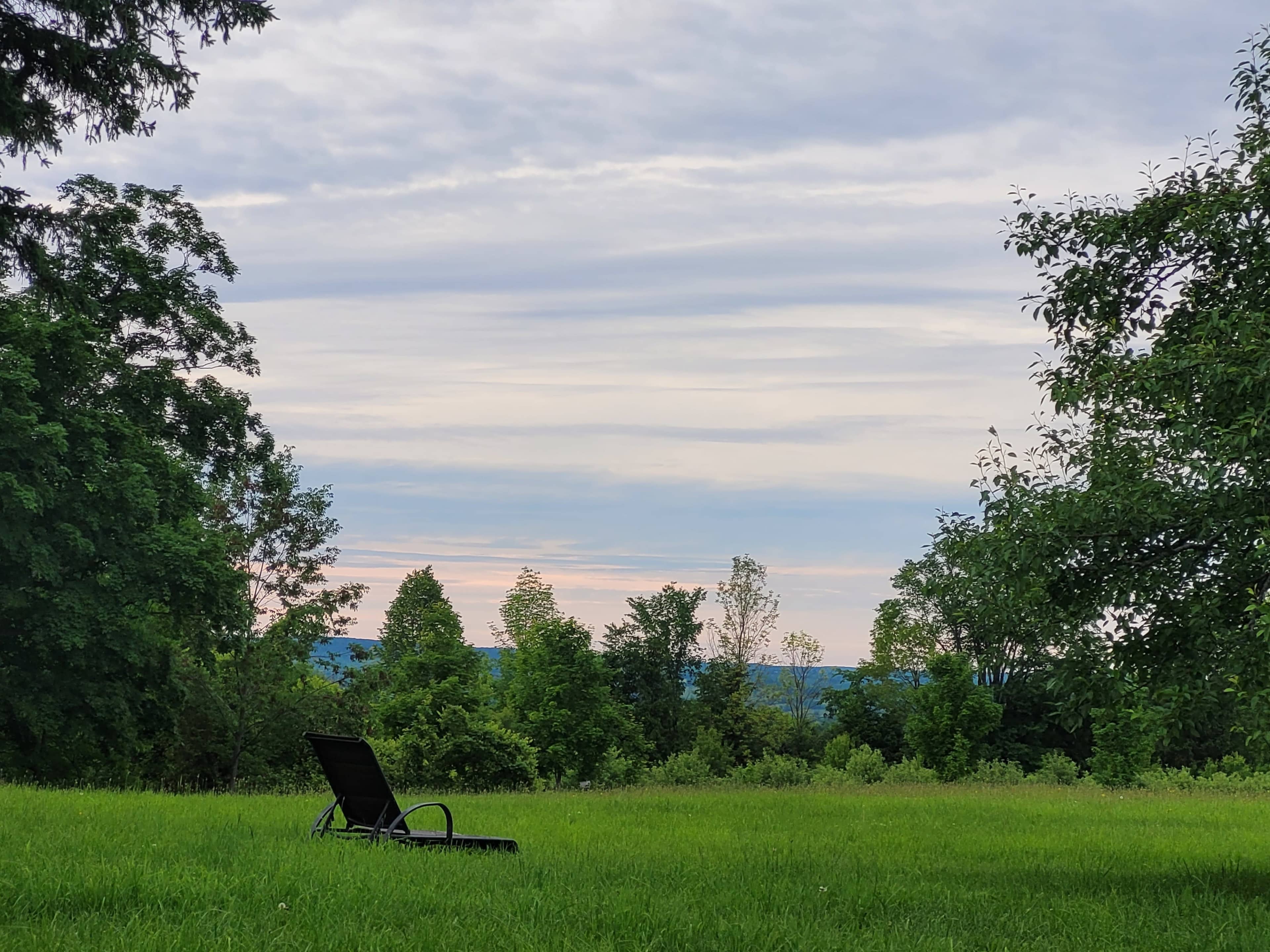 A lone chair sits on a grassy field surrounded by trees, with a cloudy sky and distant hills in the background.