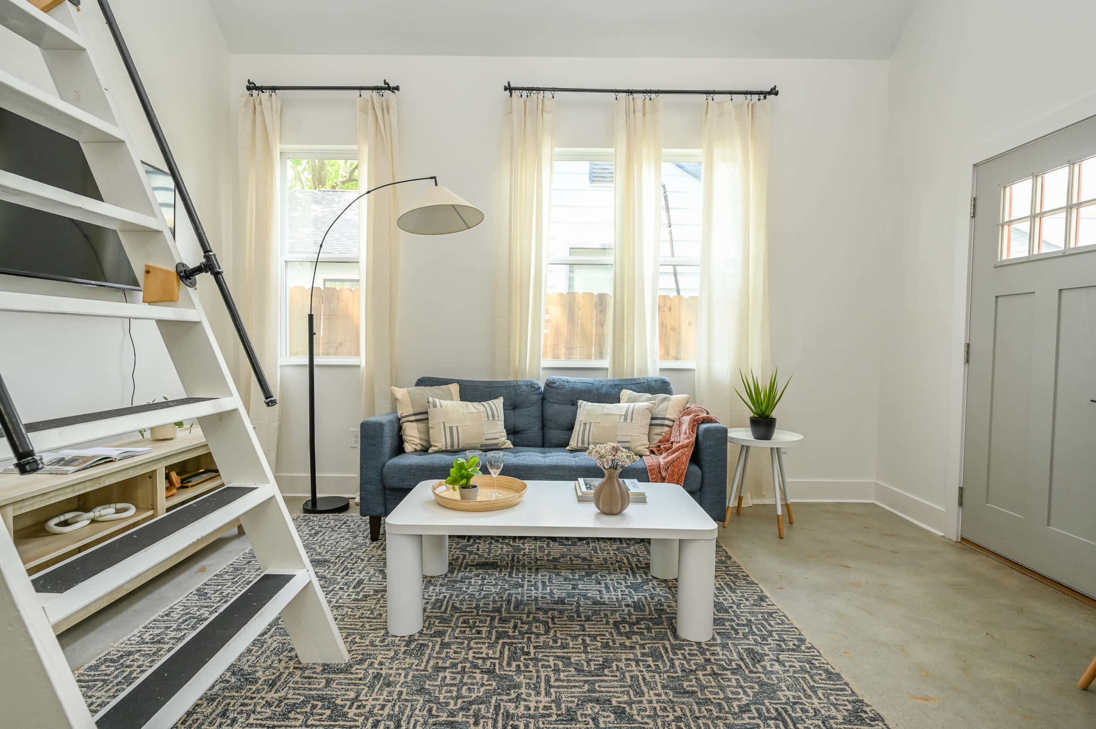 The image shows a cozy living room with a blue sofa, a white coffee table, and a staircase leading to a loft area, all illuminated by natural light from large windows.