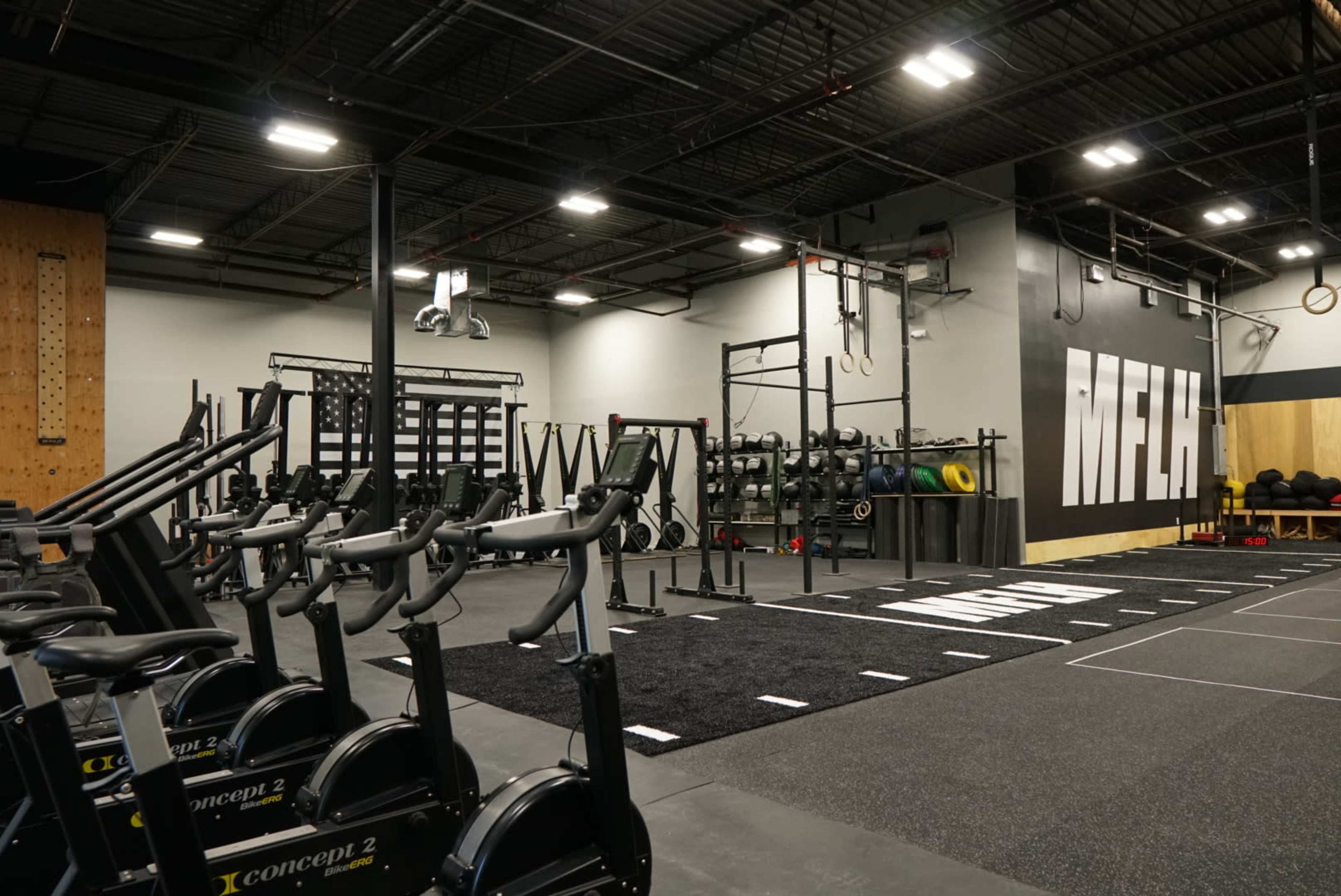 The image shows an indoor gym space featuring exercise equipment, including stationary bikes and weightlifting stations, with a large wall displaying the letters "MFLA."