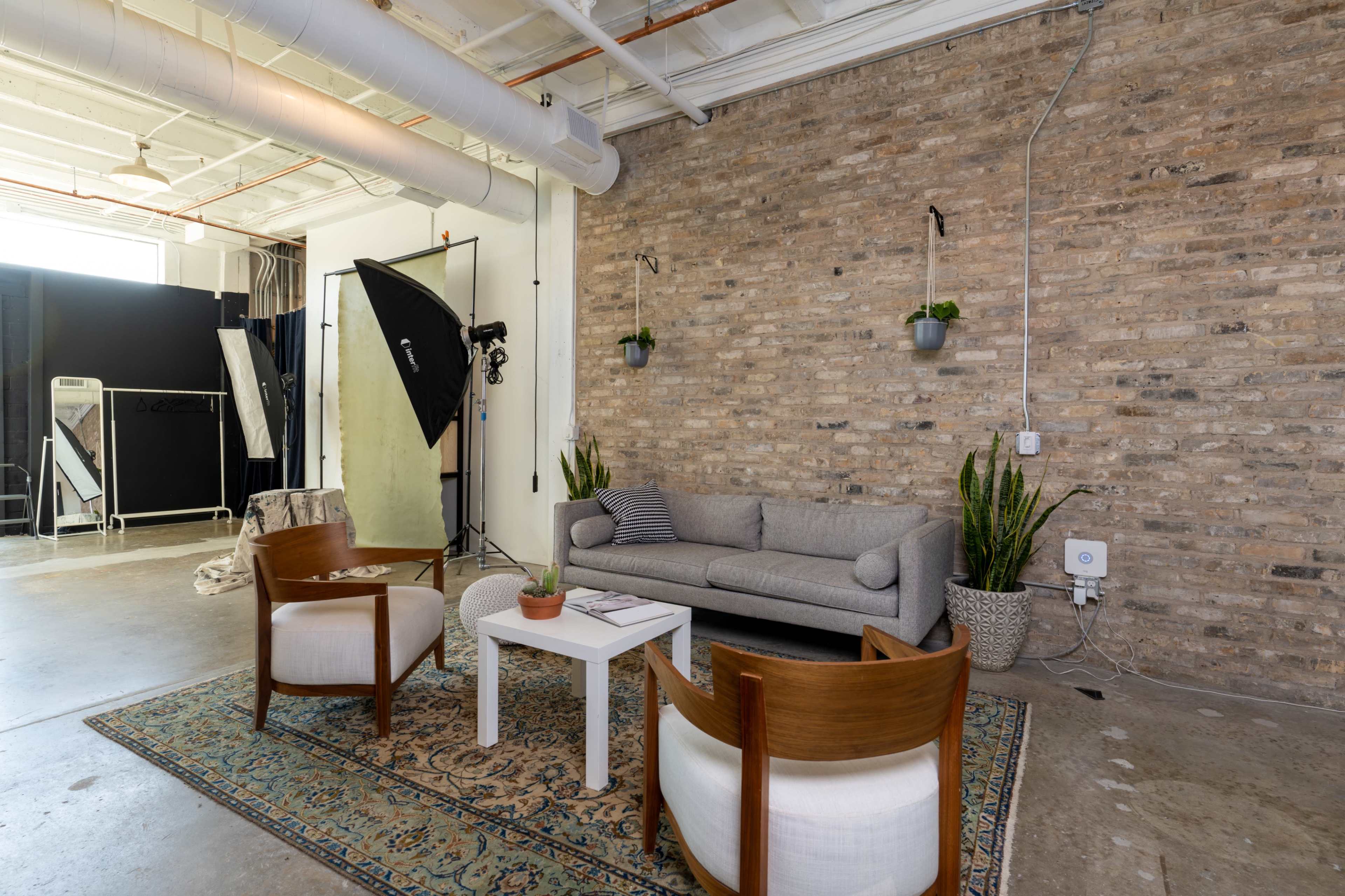 A gray sofa with white and wooden chairs is arranged around a small table on a patterned rug in a studio space with exposed brick walls.