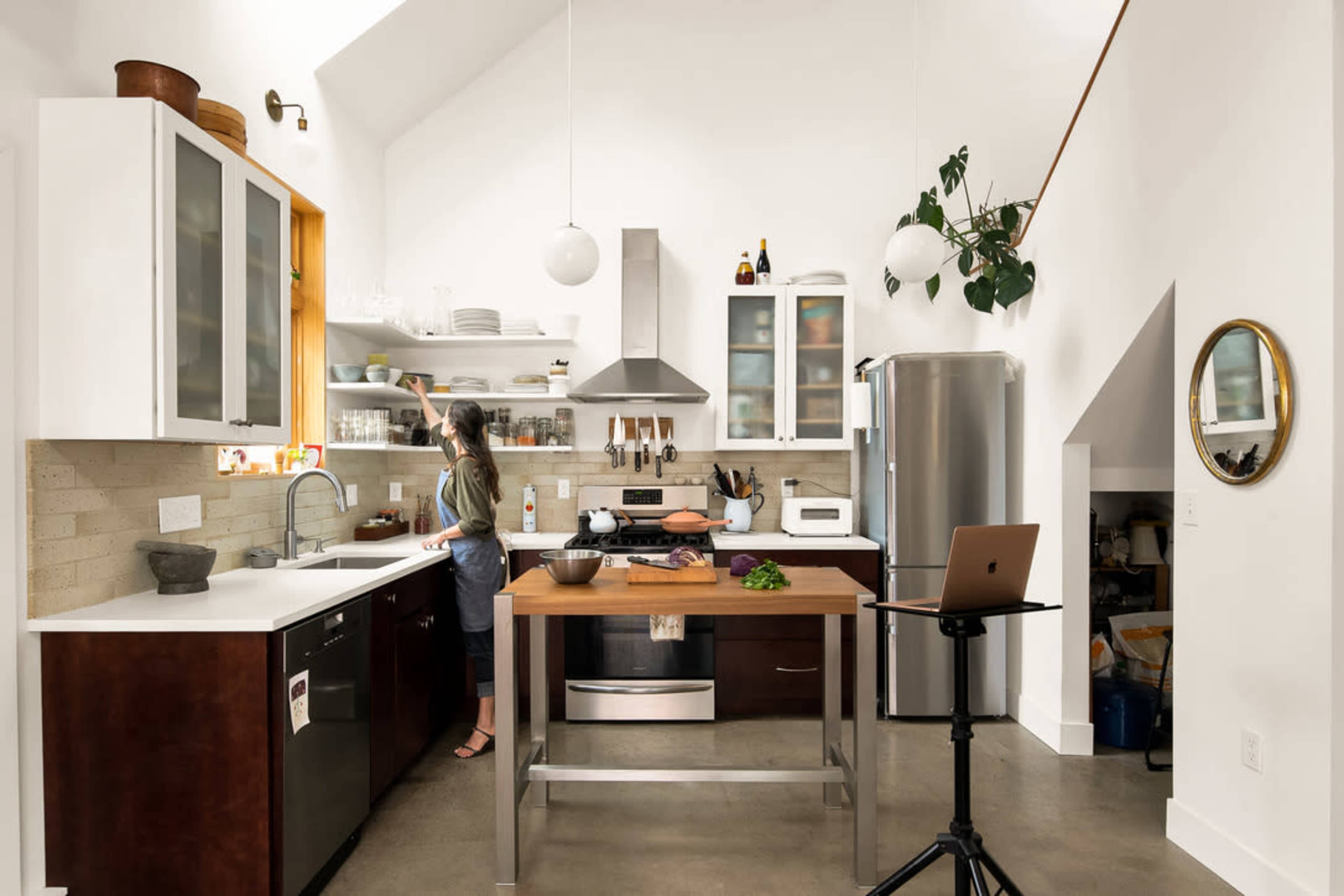 A woman stands at a modern kitchen counter, preparing food in a well-organized, brightly lit space featuring stainless steel appliances and open shelving.
