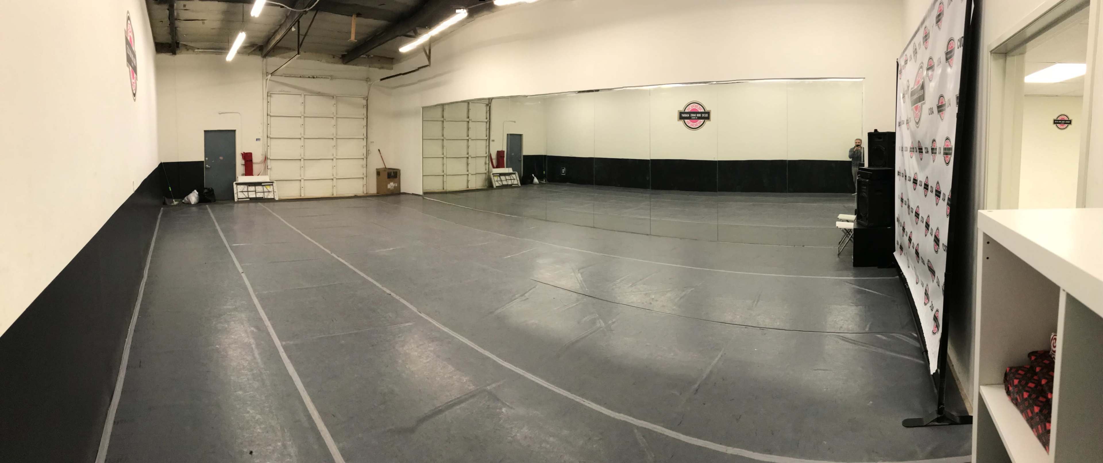 The image shows a spacious dance studio with a mirrored wall, gray flooring, and a visible backdrop with a logo.