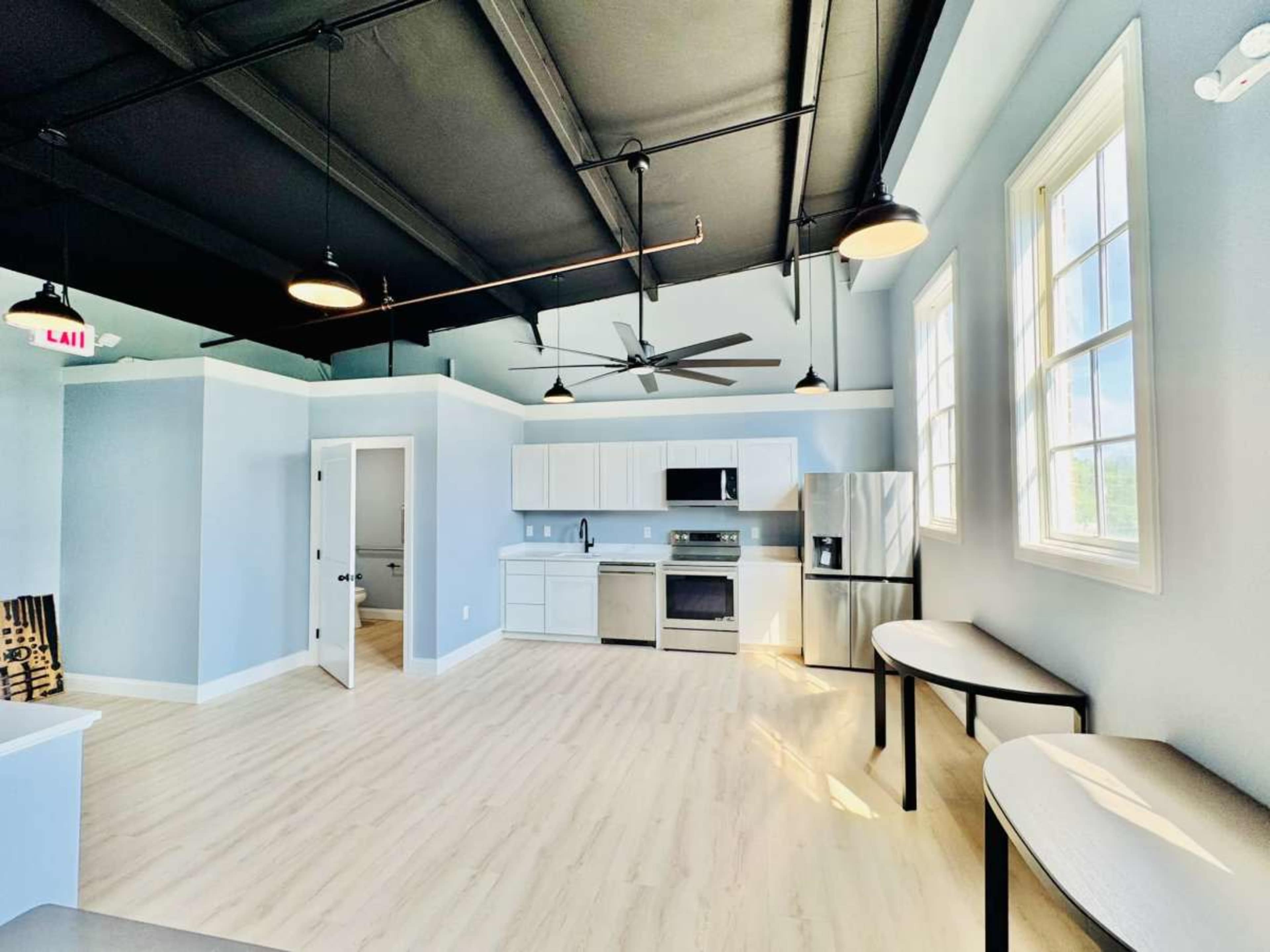 The image shows a modern, open-concept kitchen and living area with light blue walls, stainless steel appliances, and large windows.