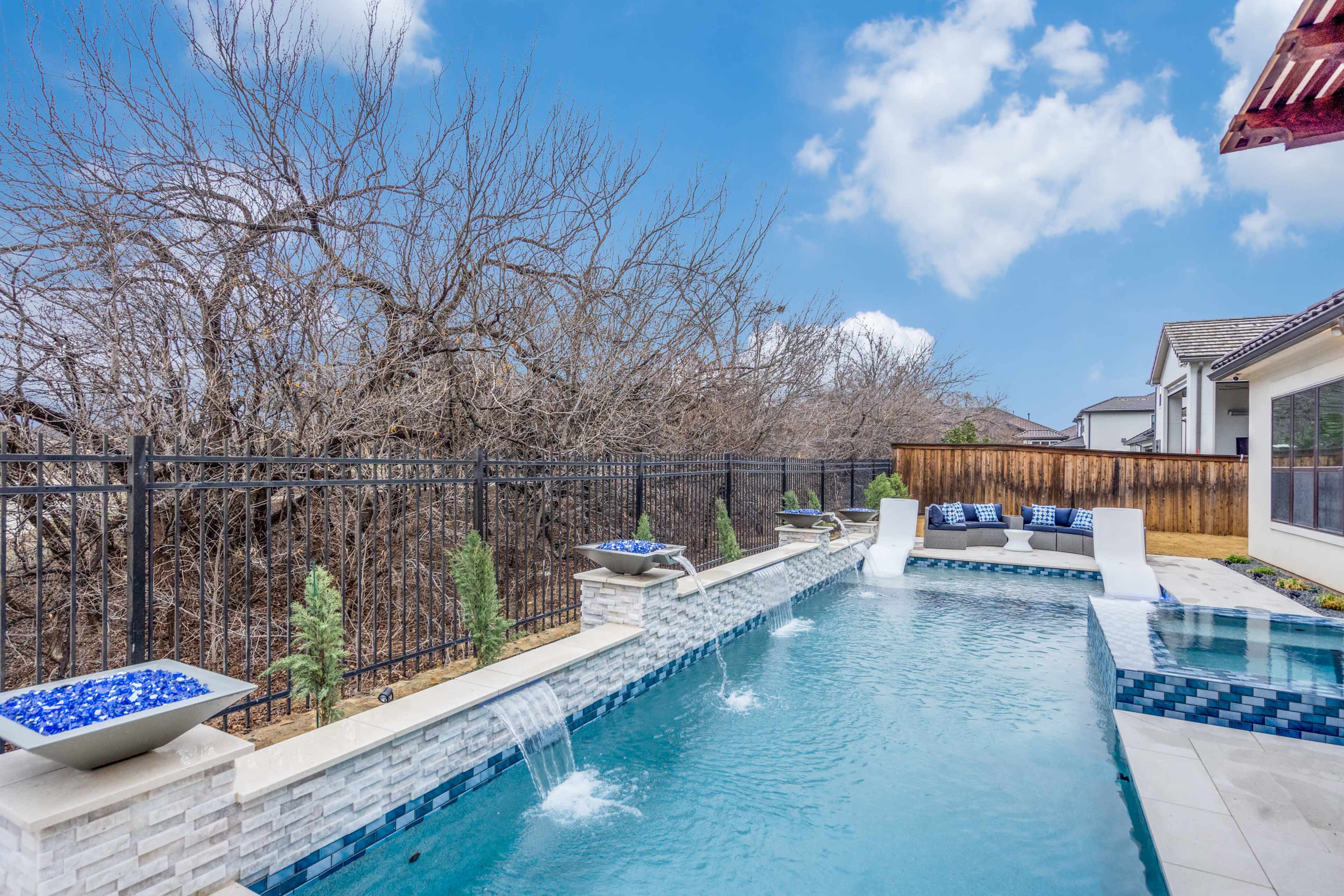 The image shows a swimming pool with water features surrounded by a fence and bare trees in the background.