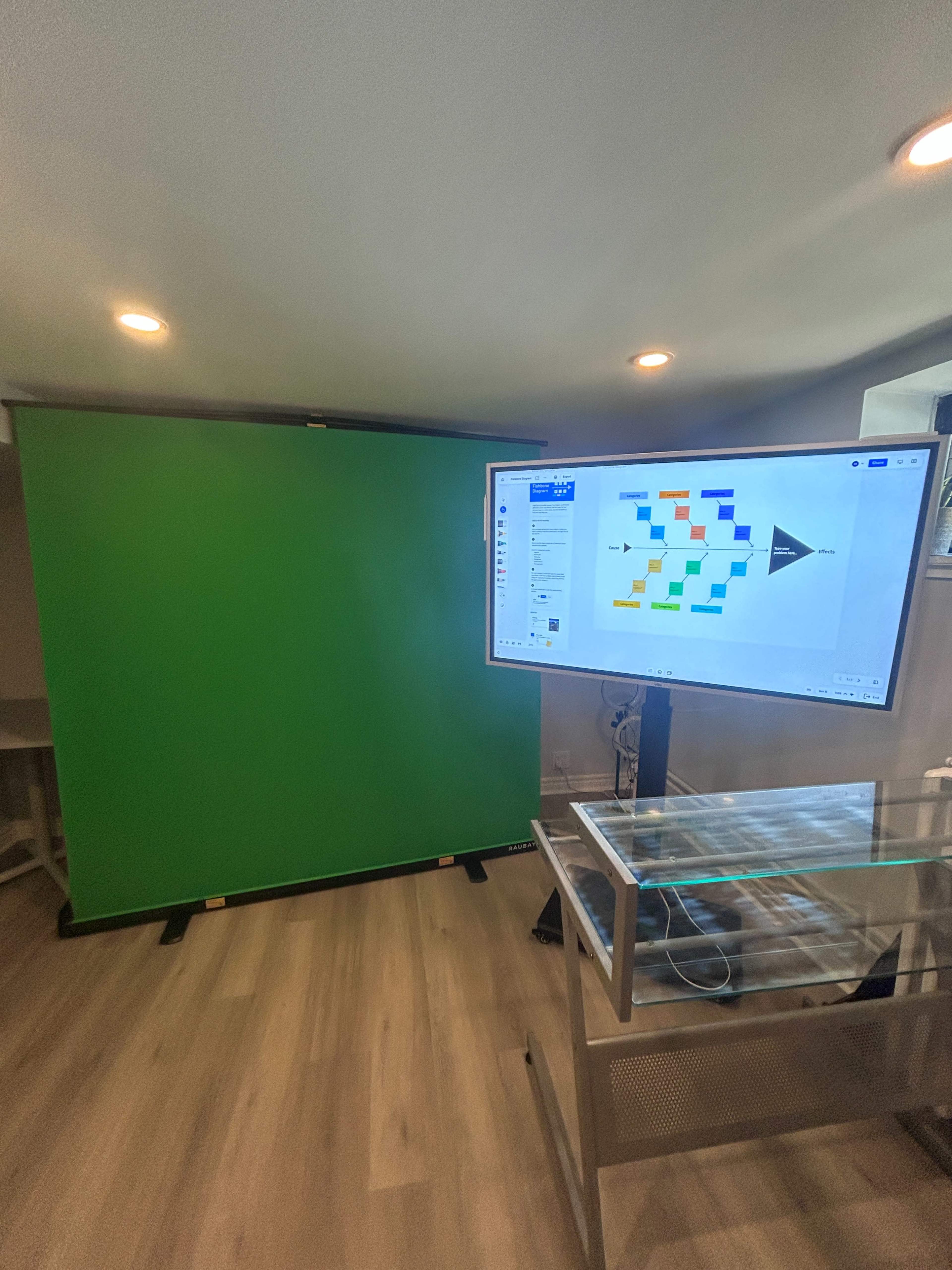 The image shows a room with a green screen setup and a large monitor displaying a colorful presentation.