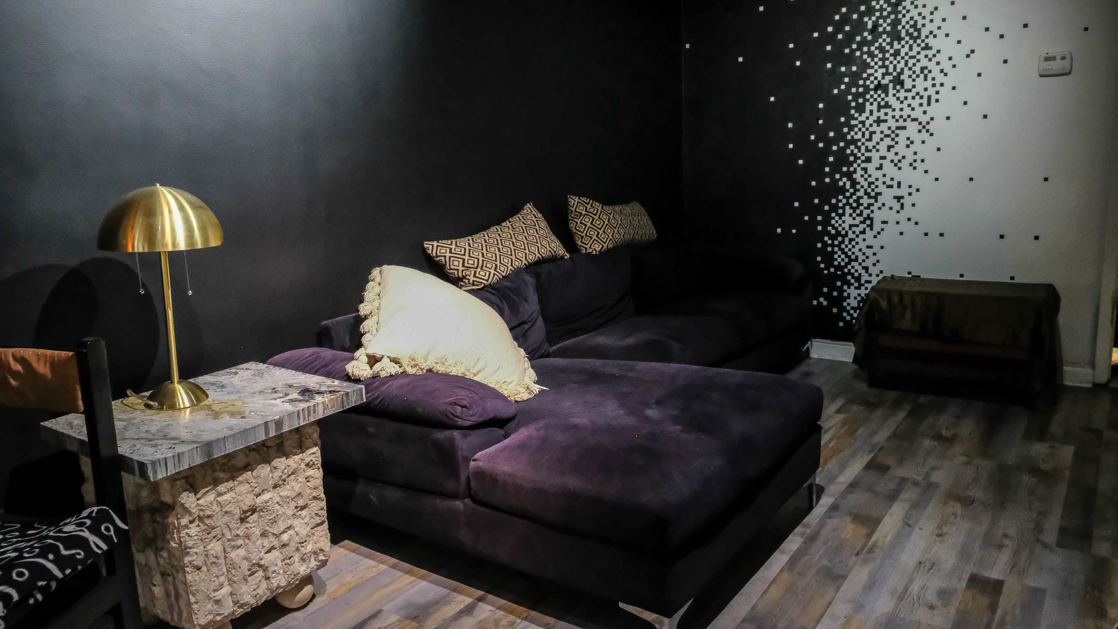The image shows a cozy lounge area featuring a dark wall, a large purple sectional sofa with decorative pillows, a small marble side table, and a gold lamp.