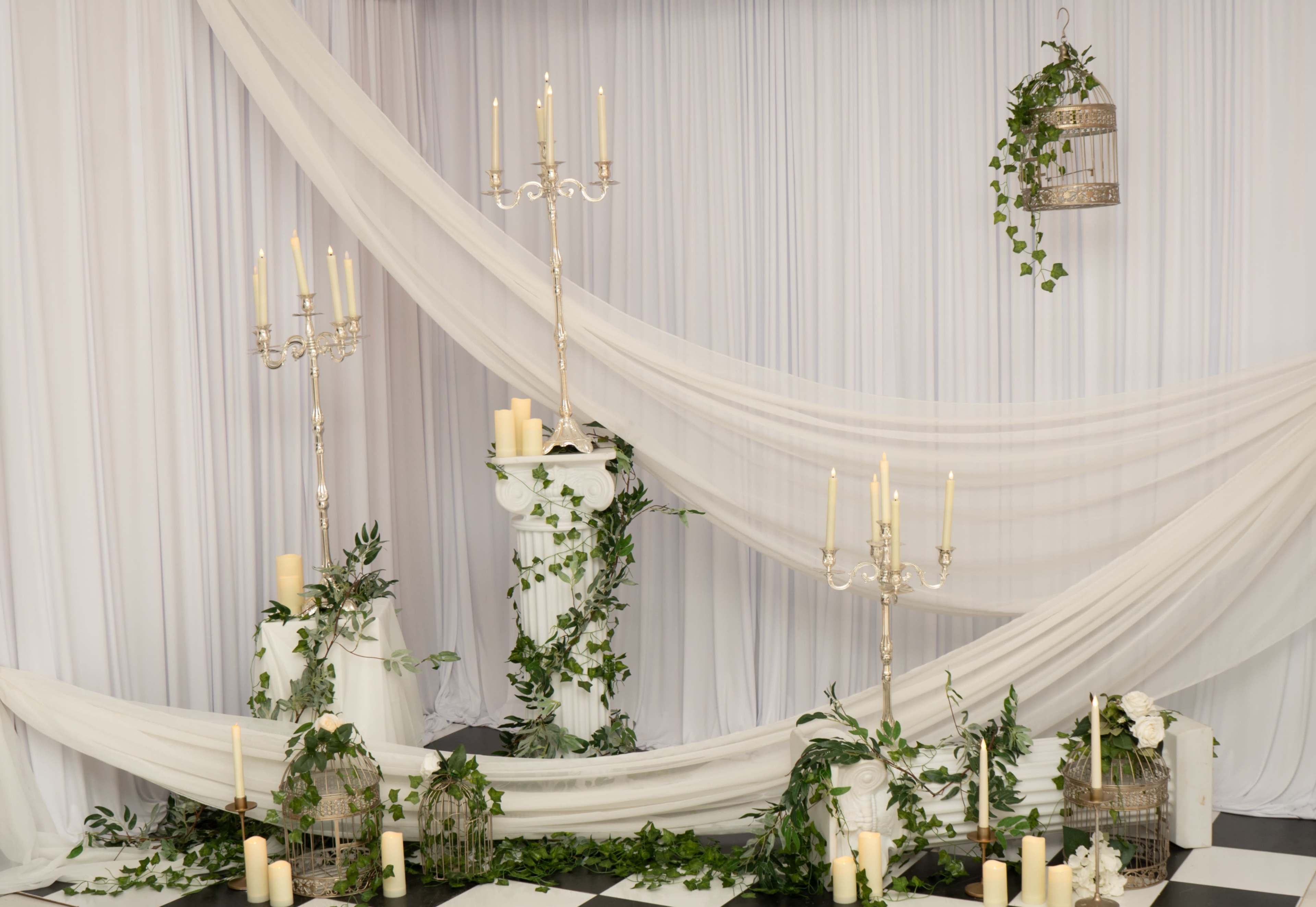 The scene features elegant white drapery, tall candle holders, and greenery arranged around decorative columns.