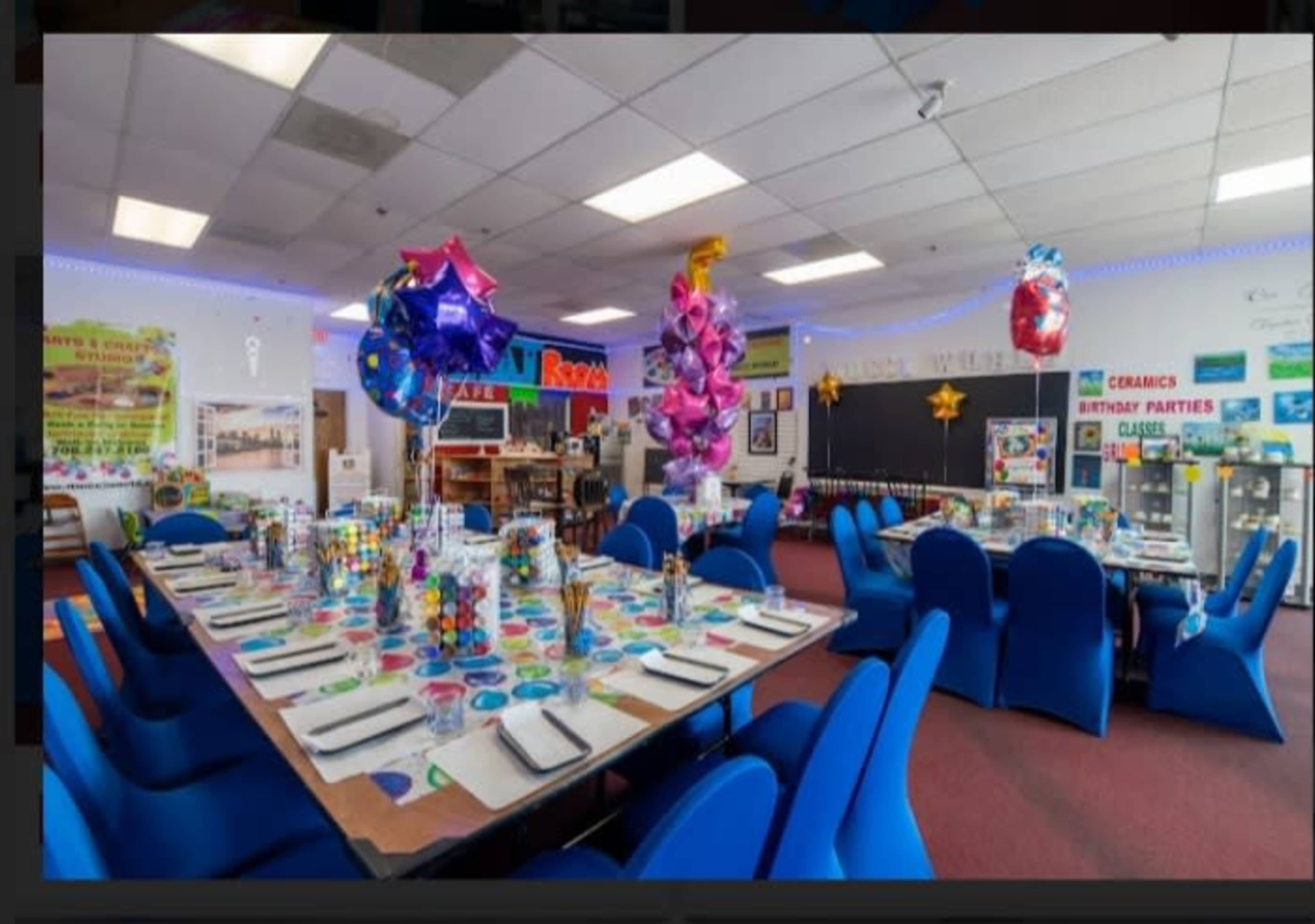 A colorful classroom decorated for a celebration, featuring a long table set with party supplies and balloons overhead.
