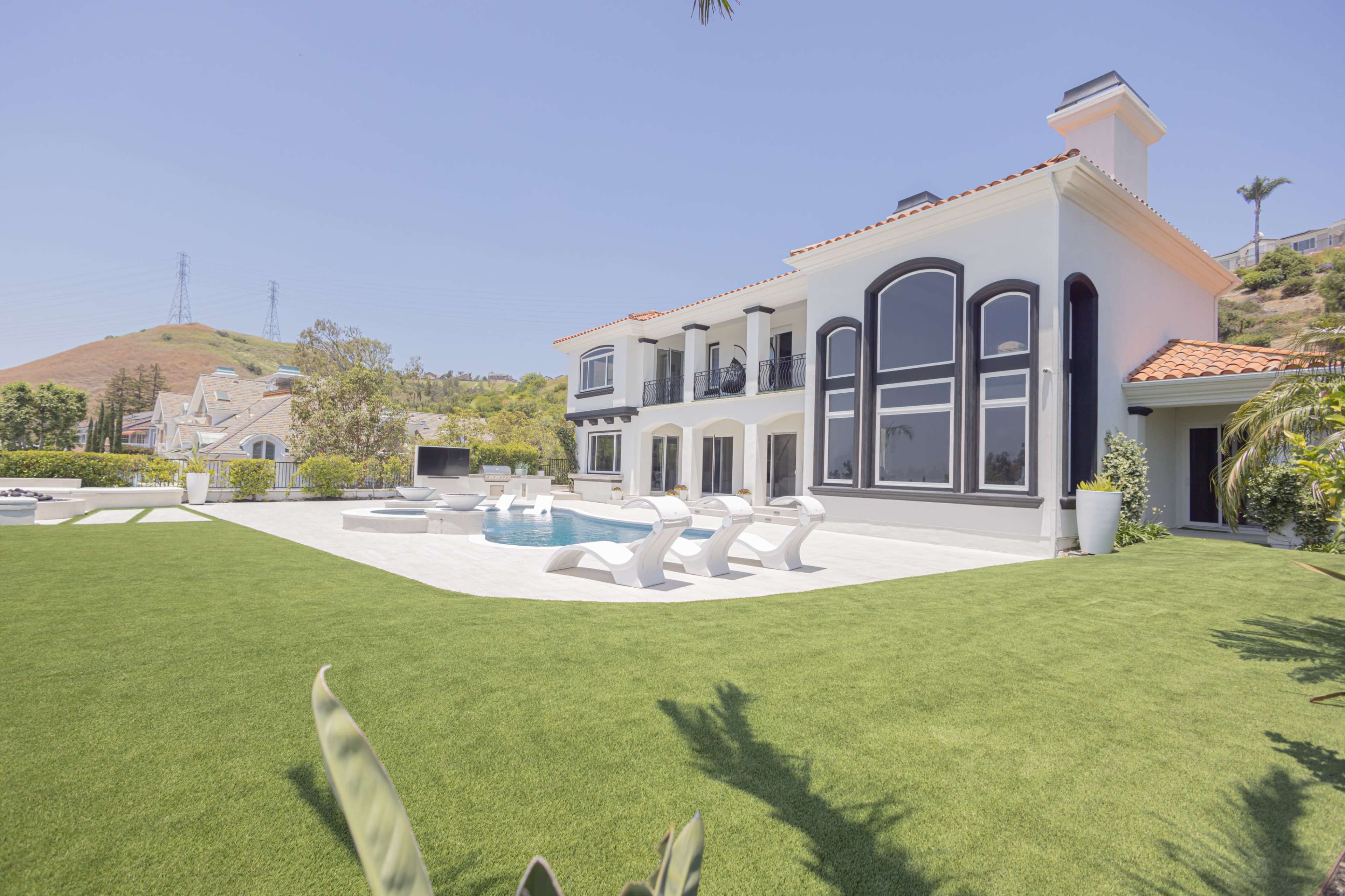 A large modern house with expansive windows and a tiled patio is surrounded by green lawn and features a swimming pool and lounge chairs.