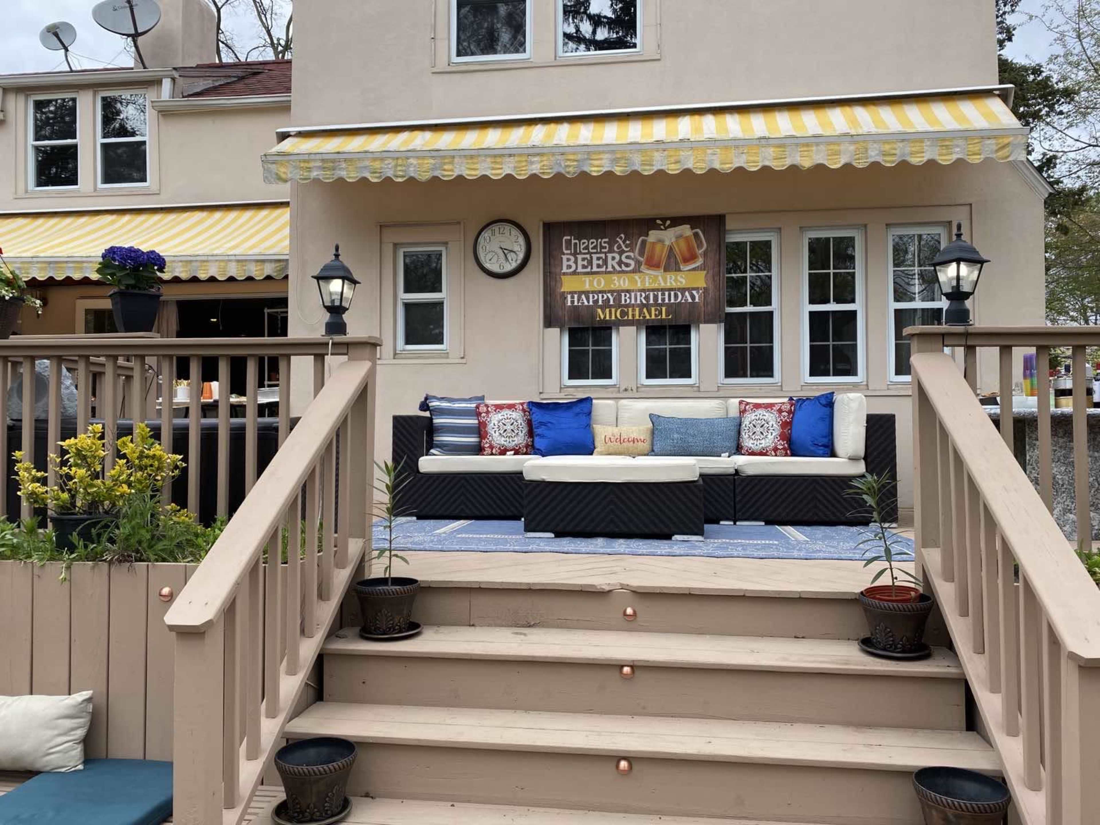 A birthday banner hangs above a patio area with a seating arrangement, flanked by flower pots and a clock on the wall of a house.