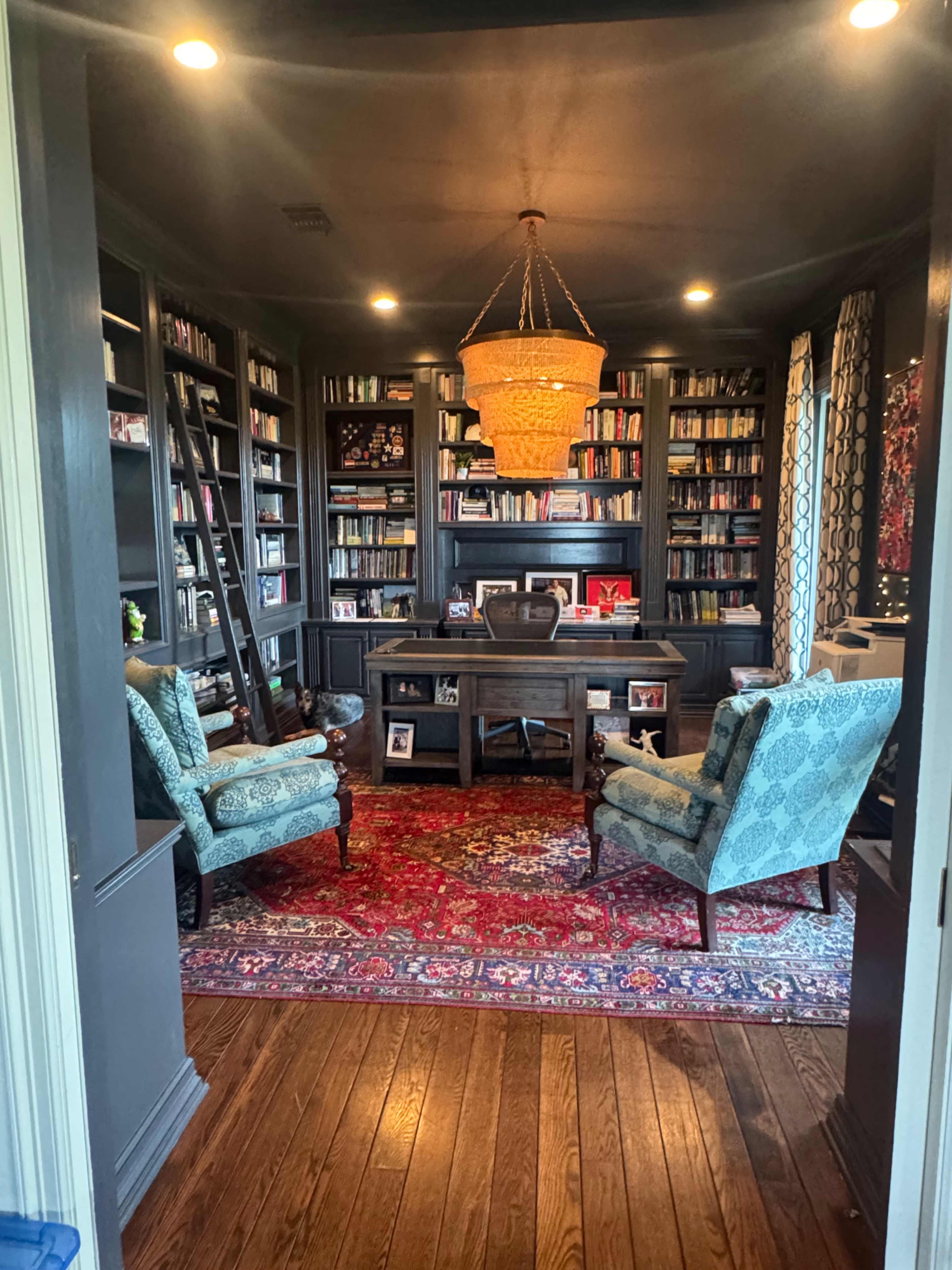 The image shows a cozy home office featuring a dark wooden desk, two patterned chairs, and shelves filled with books, illuminated by a large chandelier.