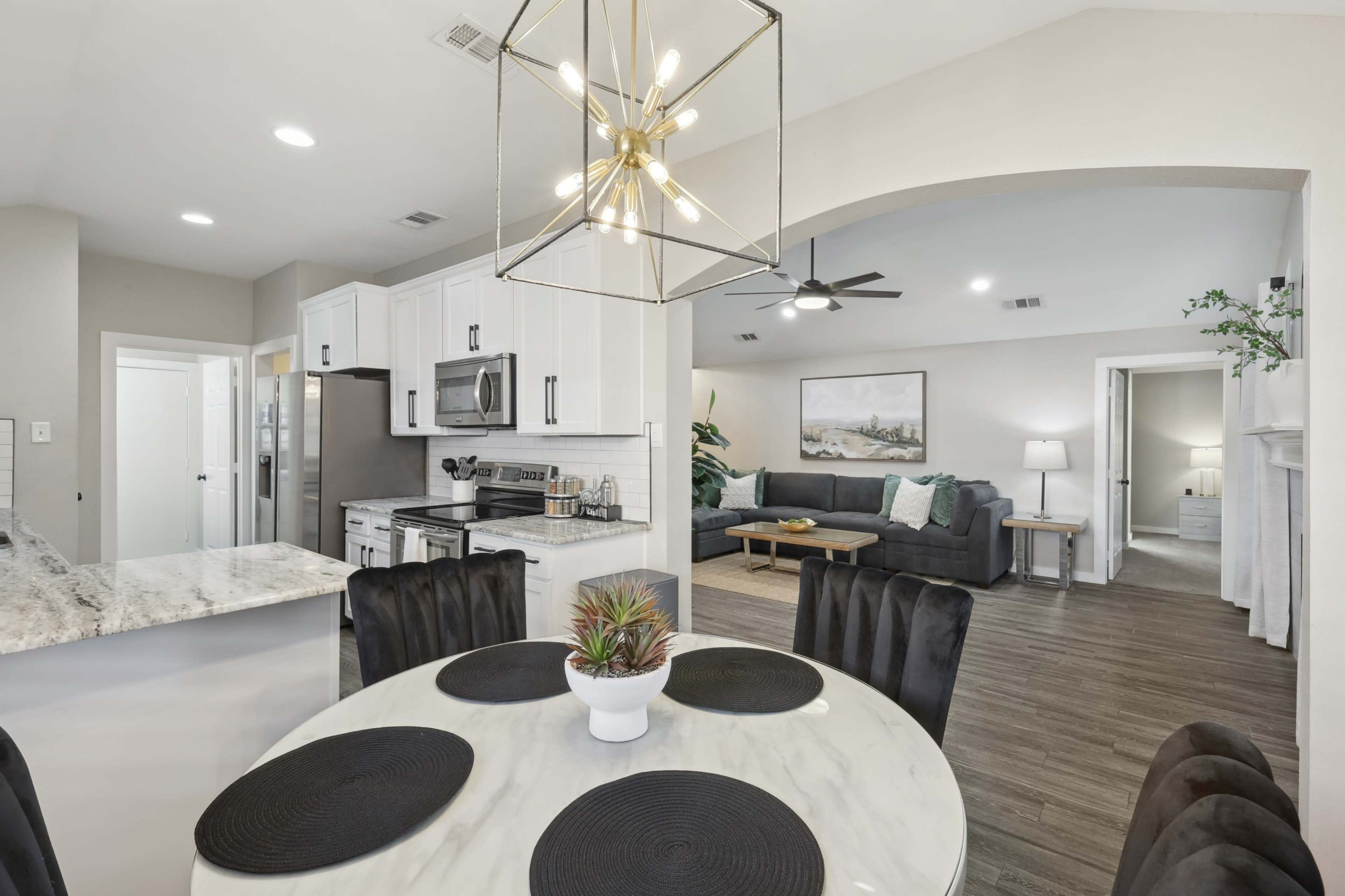 The image shows a modern dining area with a round white table and black chairs, leading into a spacious living room with contemporary furniture and a light-colored wall decor.