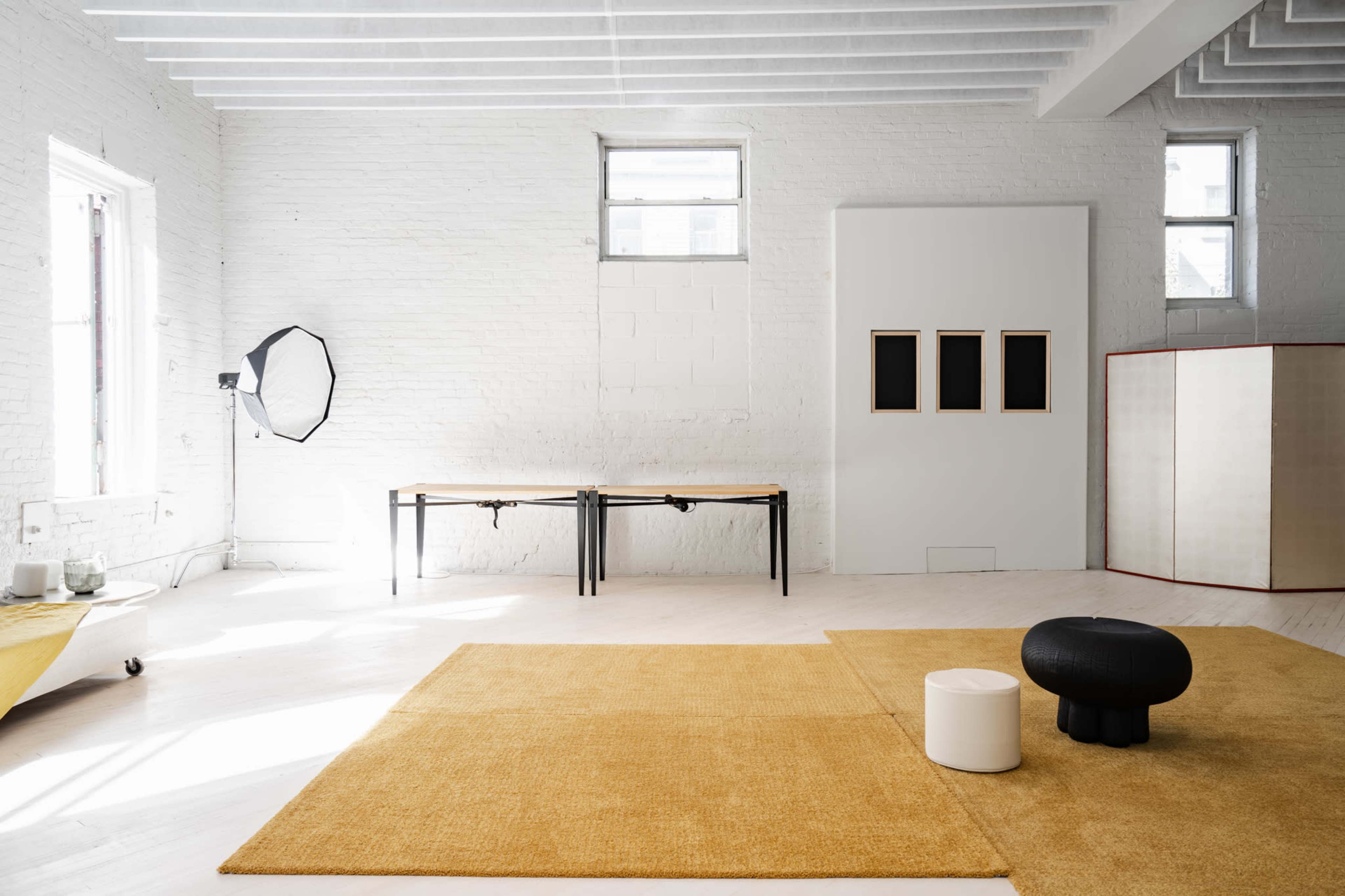 The image shows a spacious, minimalist room with a wooden table, two small stools, a large yellow rug, and three windows allowing natural light.