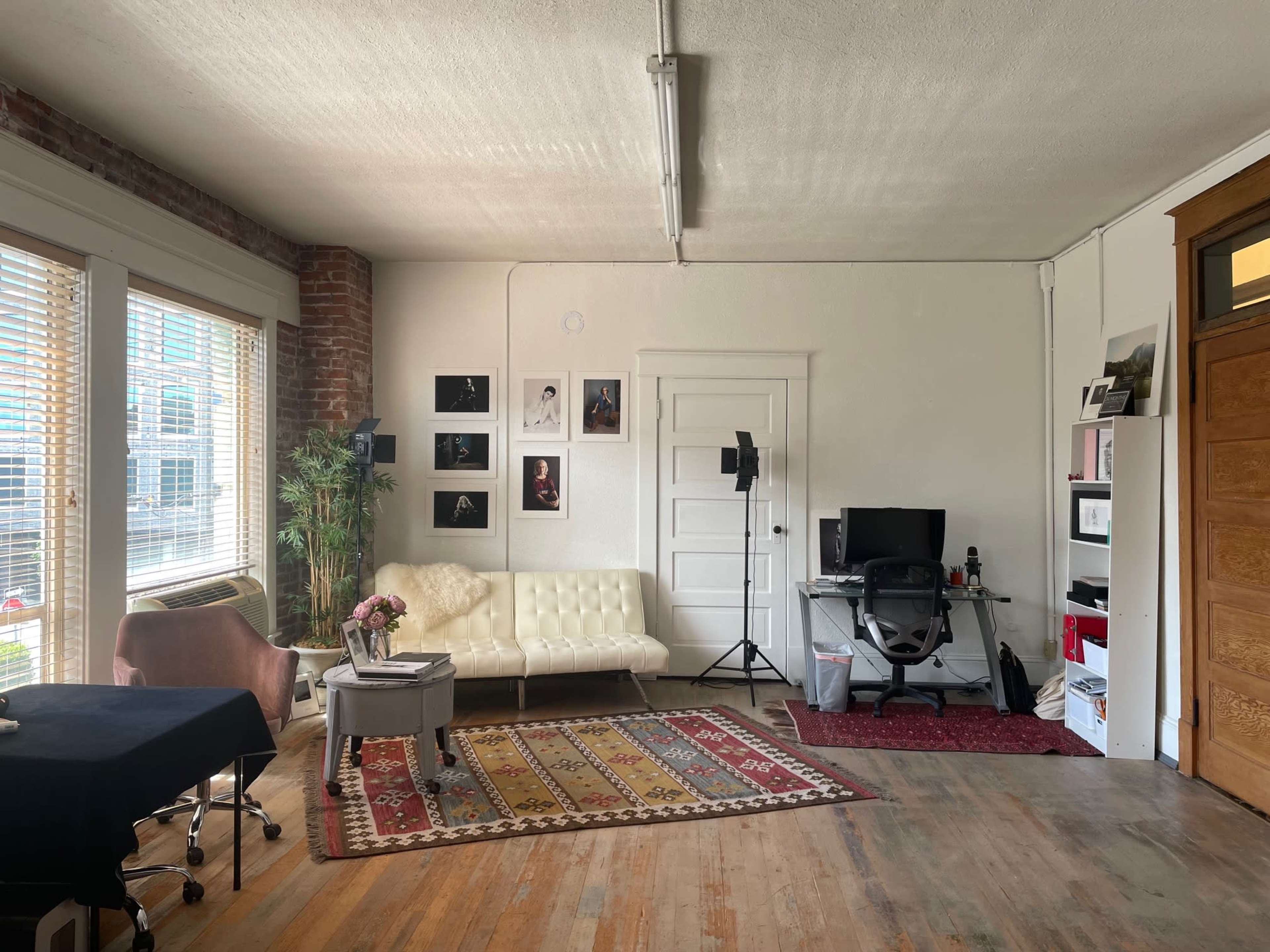 A bright office space featuring a white sofa, a desk with a computer, framed photographs on the wall, and a large window allowing natural light to enter.