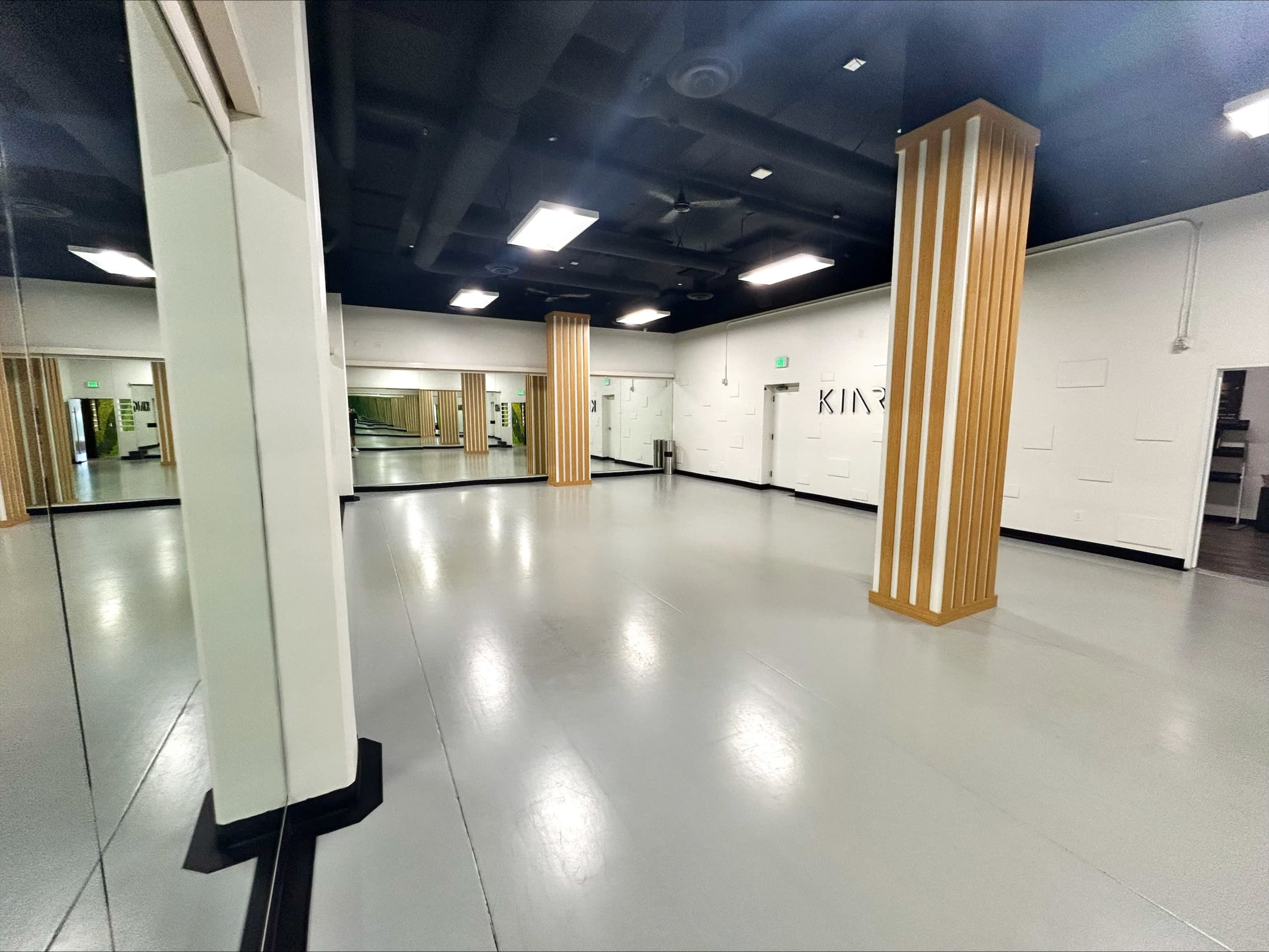 The image shows a spacious, empty studio with smooth grey flooring, white walls, and tall wooden columns.