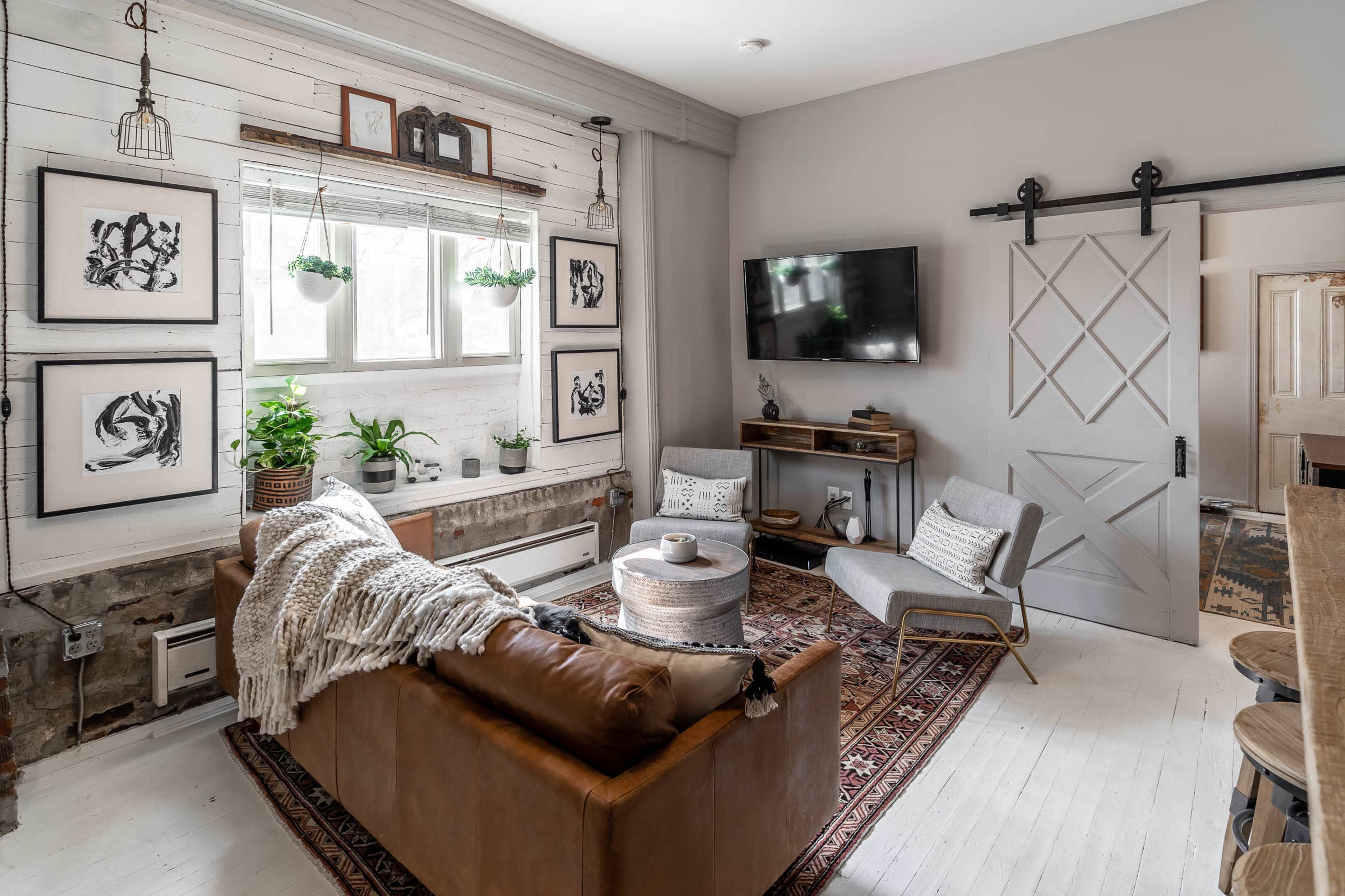 A cozy living room with a leather sofa, a round coffee table, framed artwork on the walls, and a large window surrounded by indoor plants.