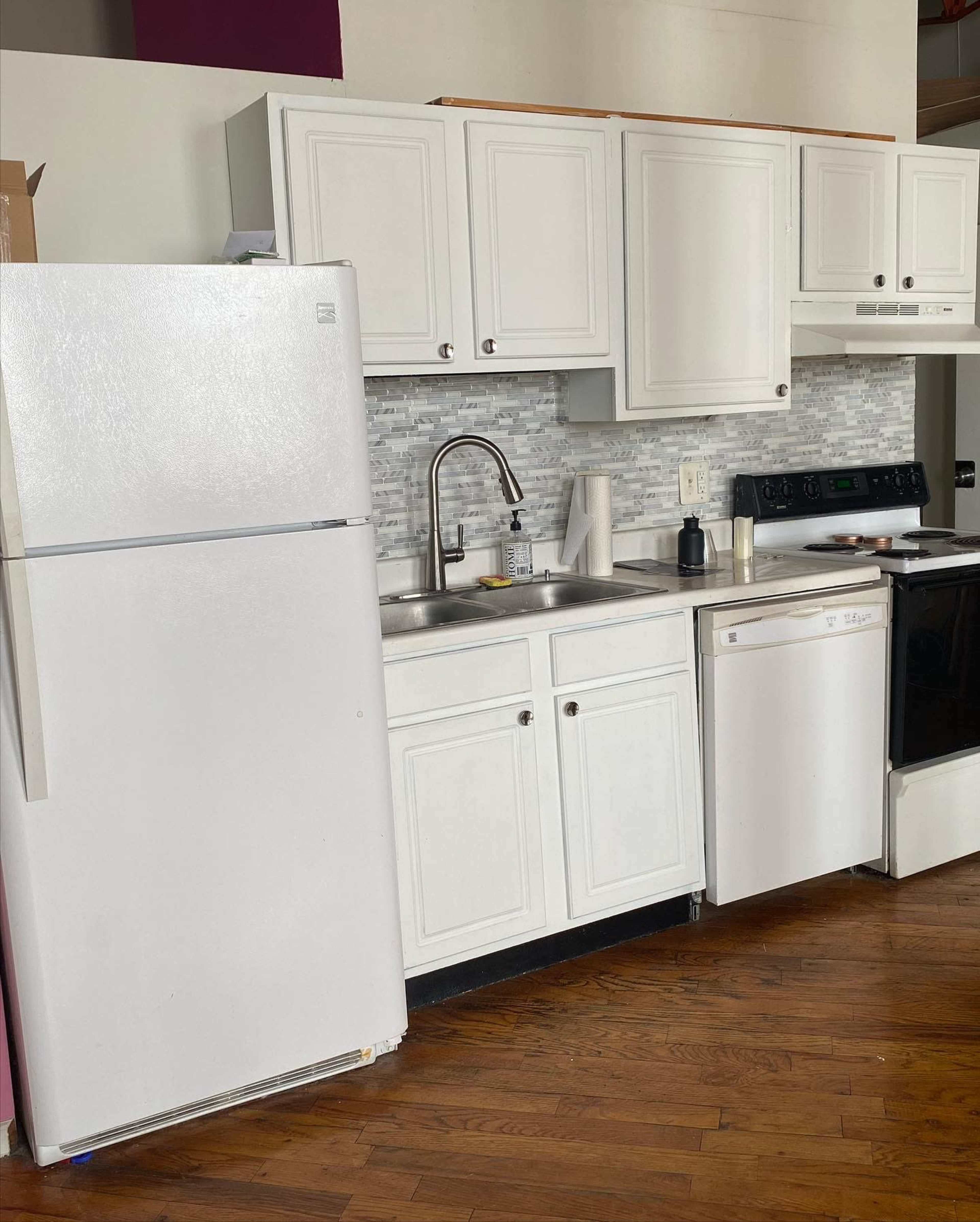 A kitchen with white cabinets, a stainless steel sink, a refrigerator, a dishwasher, and a black stove, all set against a wooden floor.