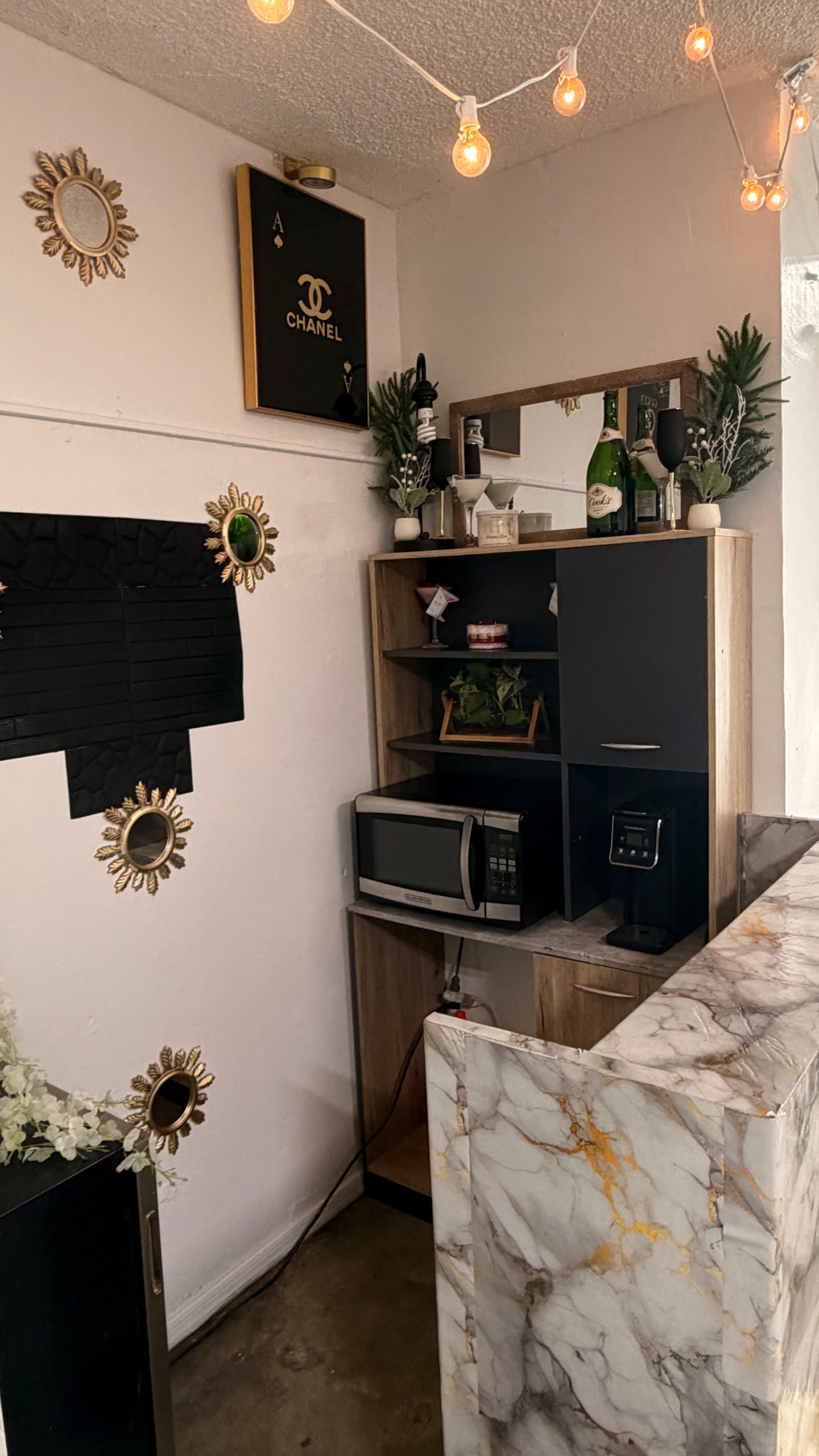 The image shows a small, stylish kitchenette area featuring a microwave, a beverage cooler, decorative plants, and a marble countertop, with lights hanging above.