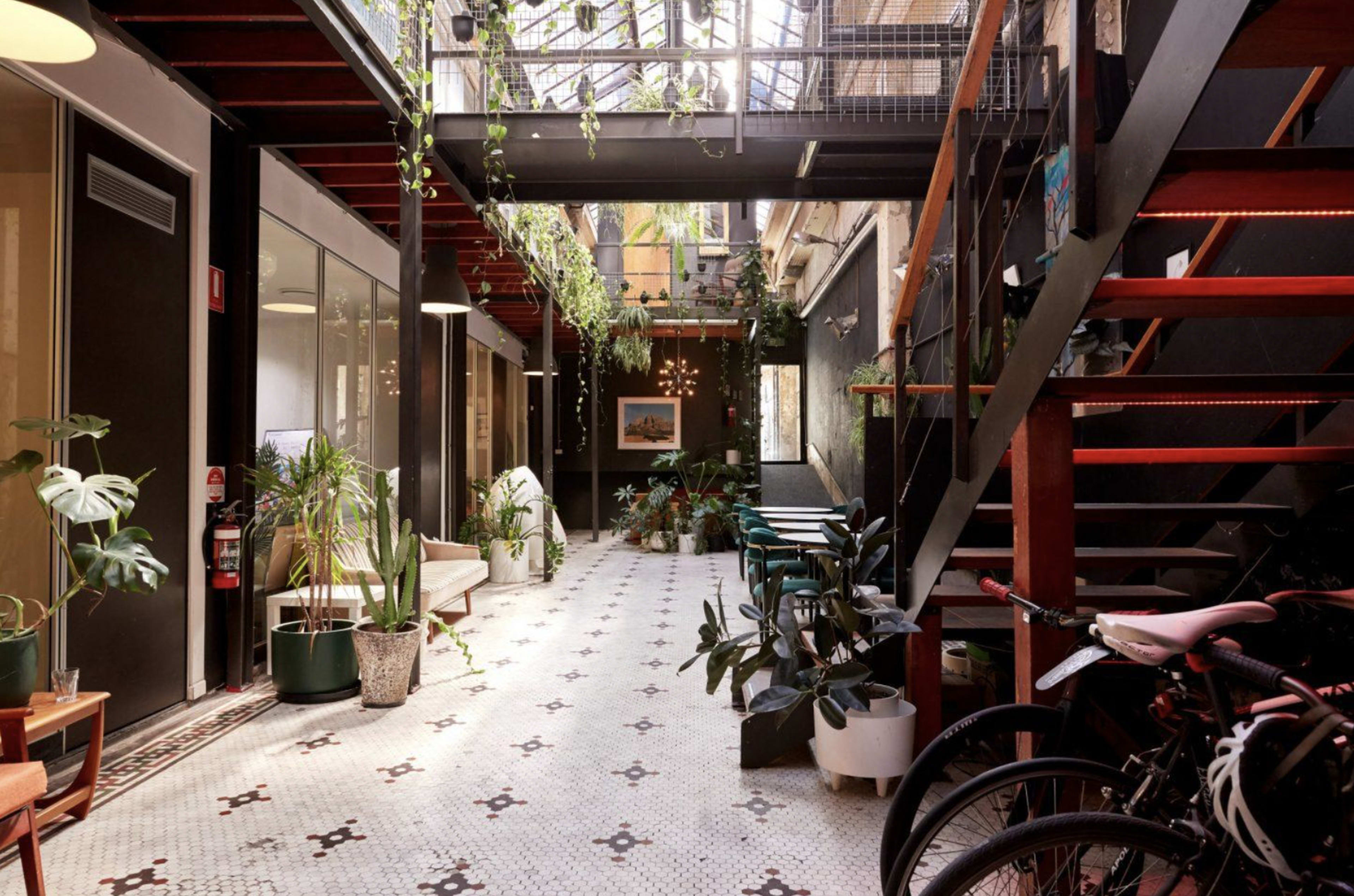The image shows a spacious, indoor corridor with decorative plants, tiled flooring, and bicycle parking.