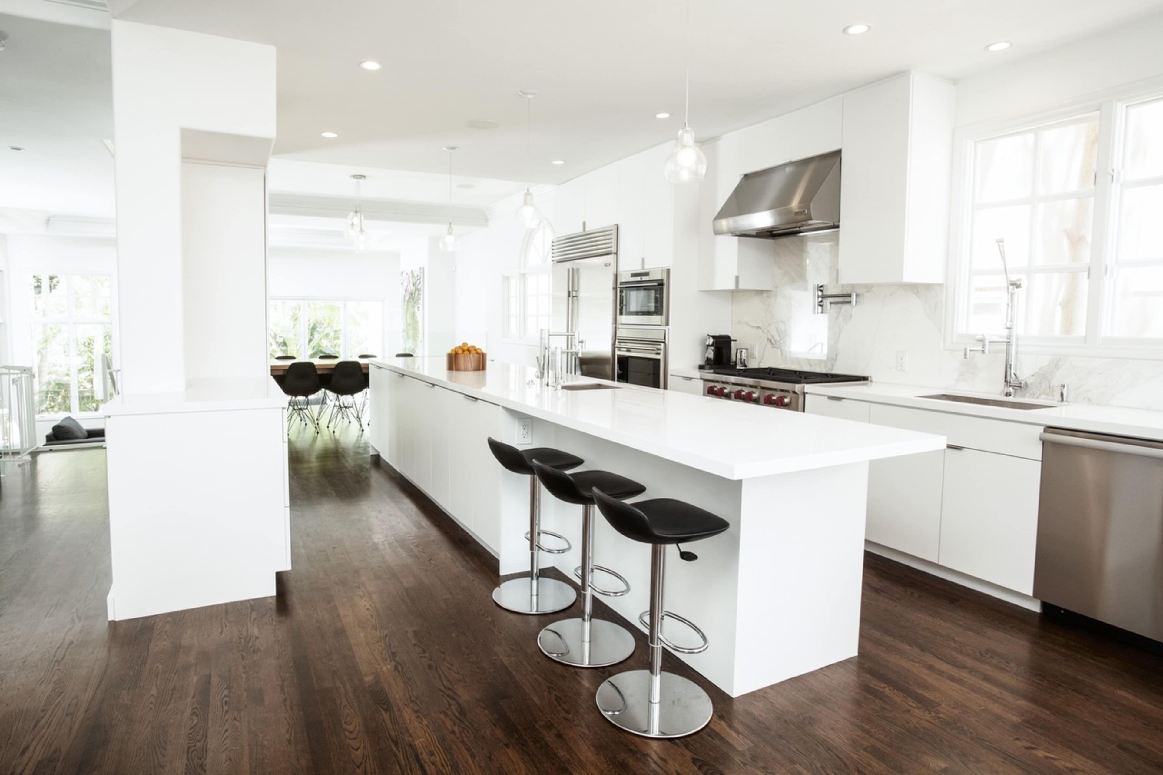 A modern kitchen features a white island counter with black bar stools, stainless steel appliances, and large windows allowing natural light to flood the space.