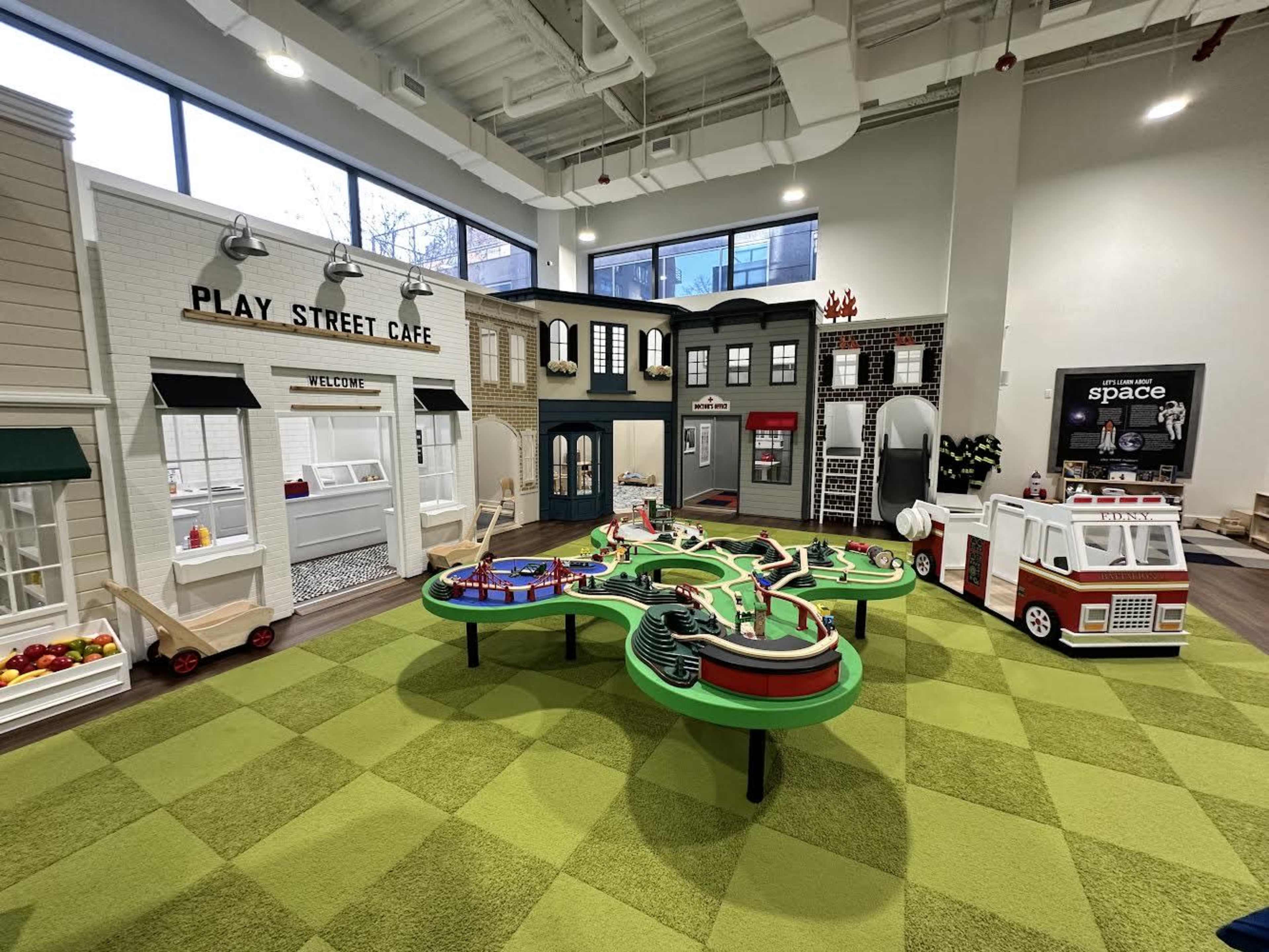 A colorful indoor play area featuring various playhouses and a miniature train set on a green carpet.