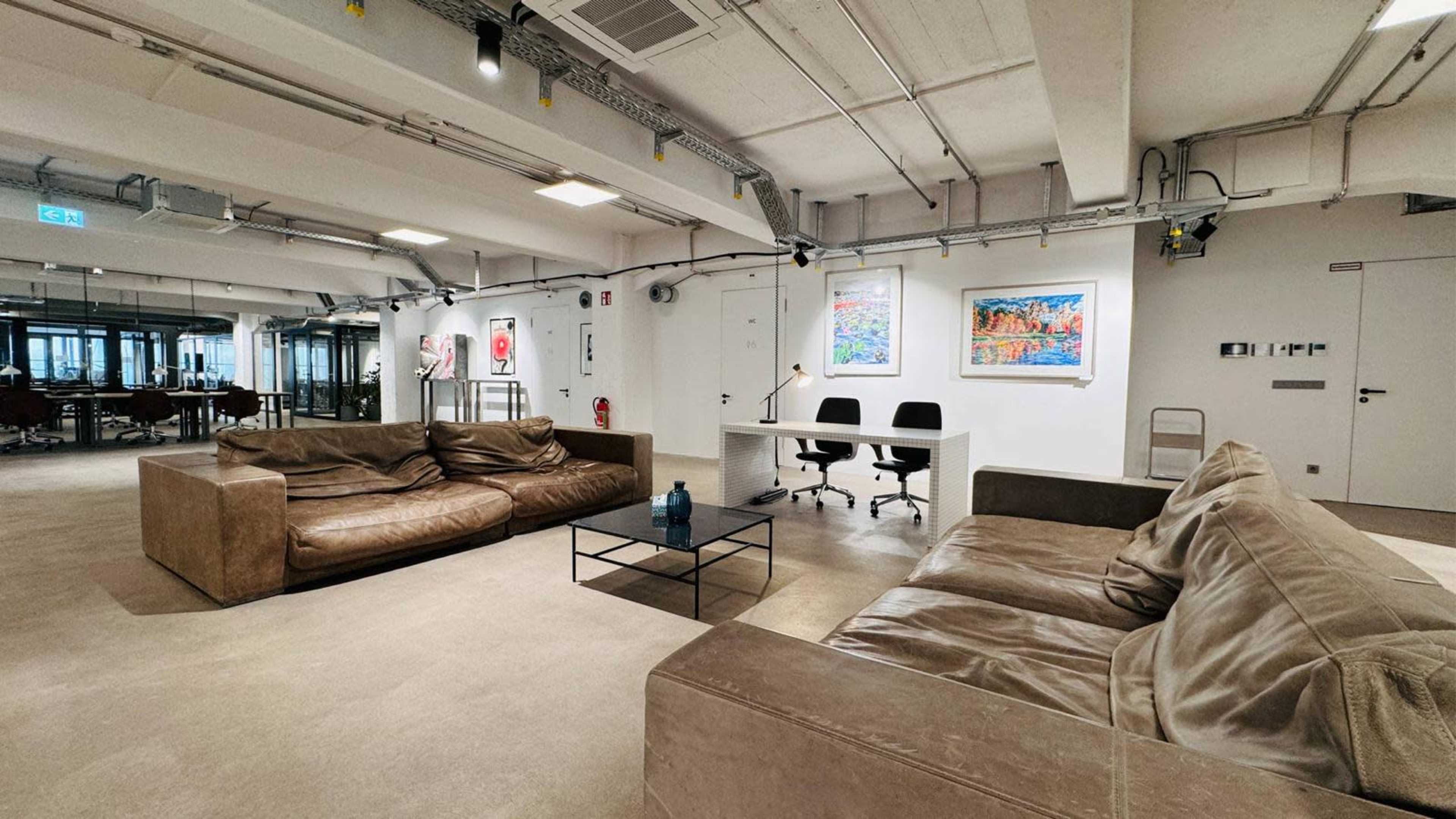 A modern office space featuring two large sofas, a coffee table, a desk with two chairs, and artwork hanging on the walls.