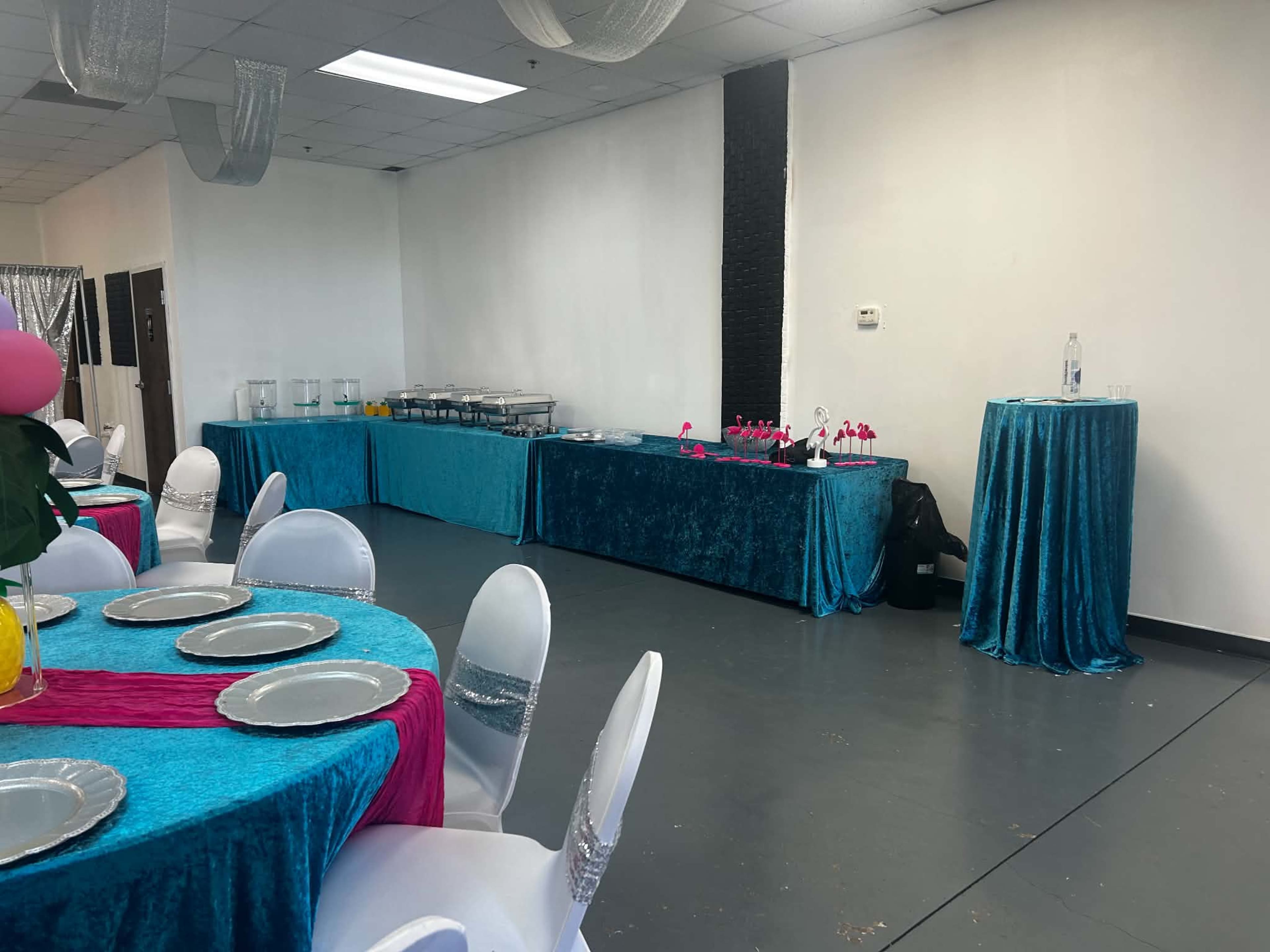 The scene shows a large room set up for an event, featuring tables covered in blue and pink tablecloths, with a buffet table and a drinks station along one wall.