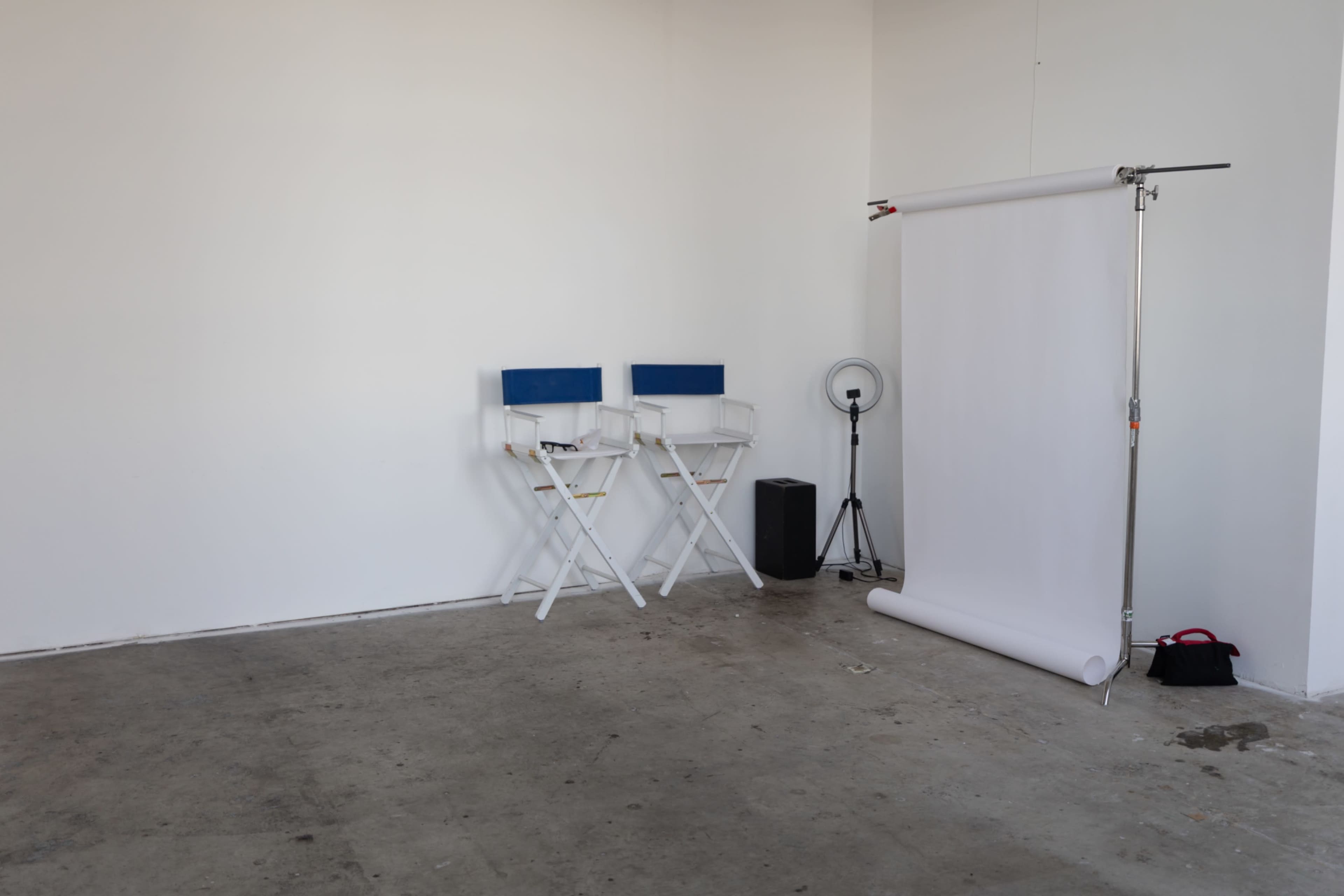 The image shows an empty studio space with two white director's chairs, a backdrop stand with a roll of white paper, a fan, and a speaker.