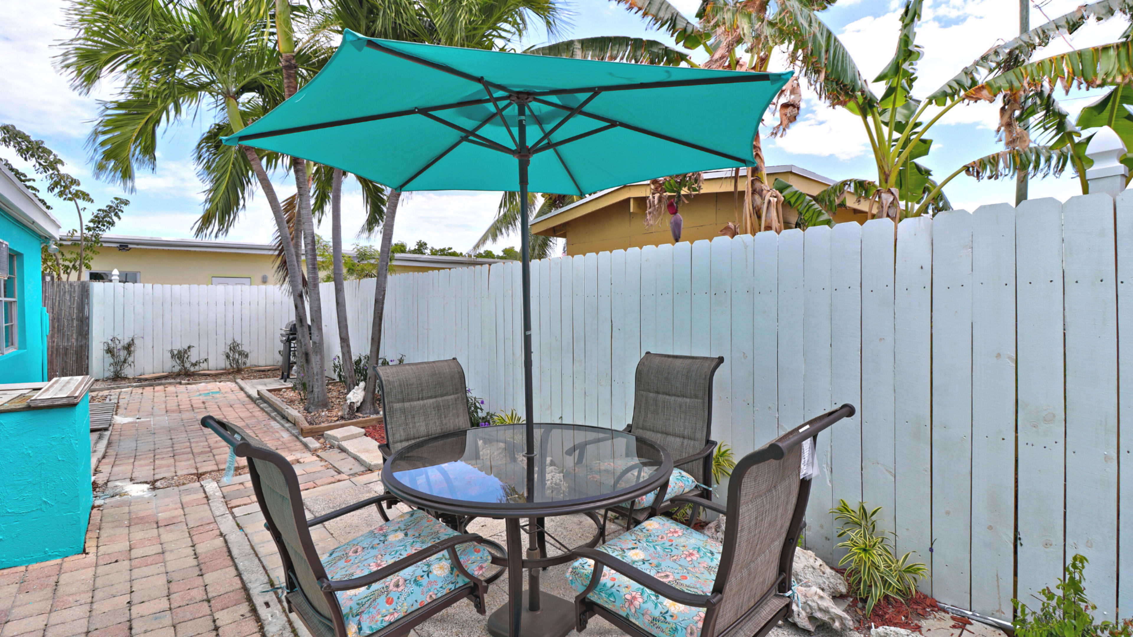 A round glass table with four chairs is shaded by a teal umbrella in a small yard surrounded by a white fence and palm trees.