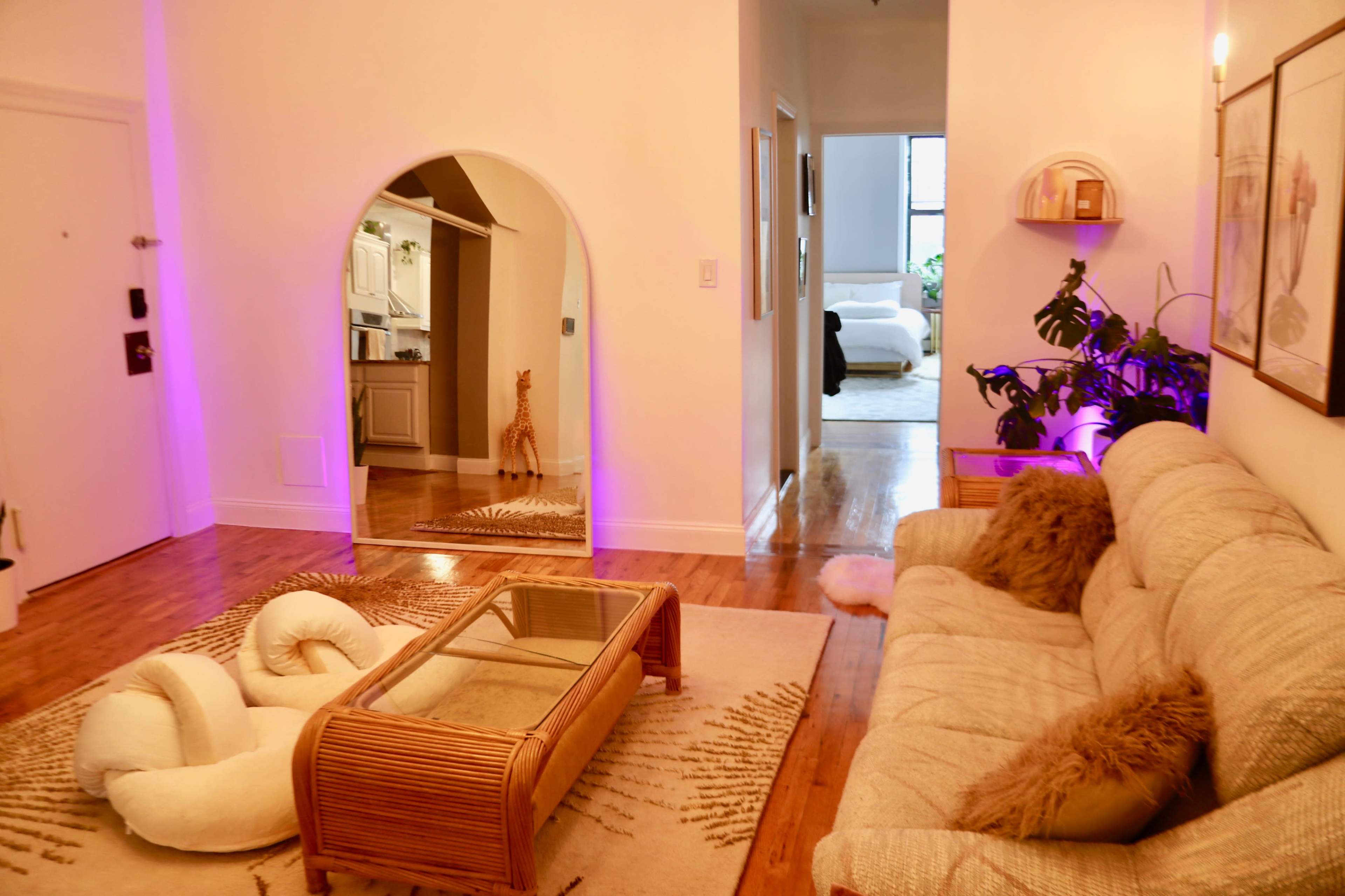 A living room with a beige sofa and a glass top coffee table, featuring a mirror with an arched frame, and a view of another room through an open doorway.